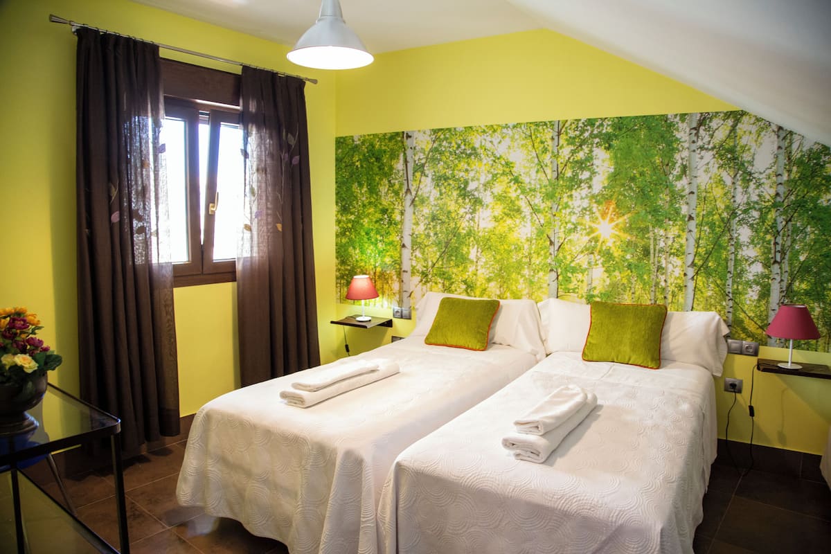 A hotel room with two beds, a large window with curtains, a wall mural of trees, and a small table with a lamp.