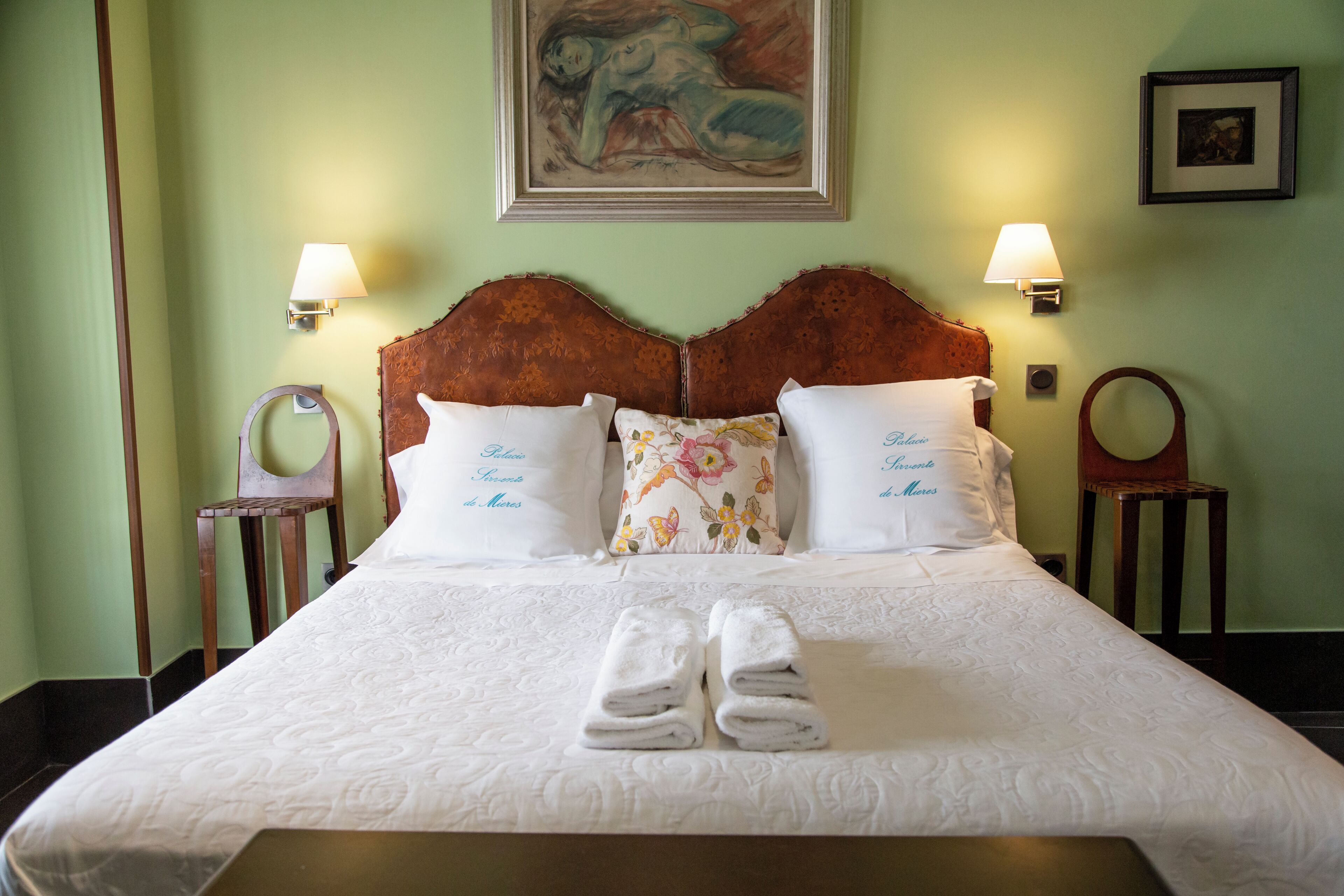 Una cama bien hecha con sábanas blancas y dos almohadas, un par de toallas dobladas y dos lámparas fijadas a la pared.