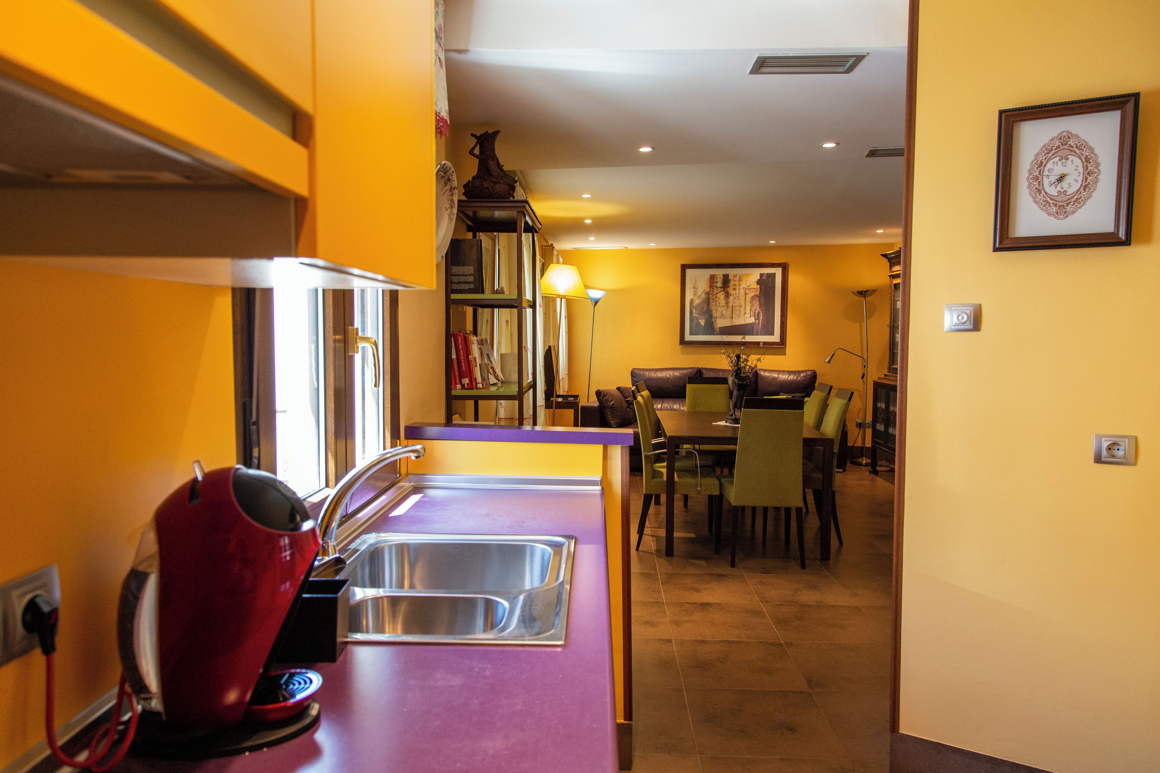 Una cocina con encimera morada, un hervidor rojo y un fregadero. Se observa un comedor con sillas y una mesa al fondo.