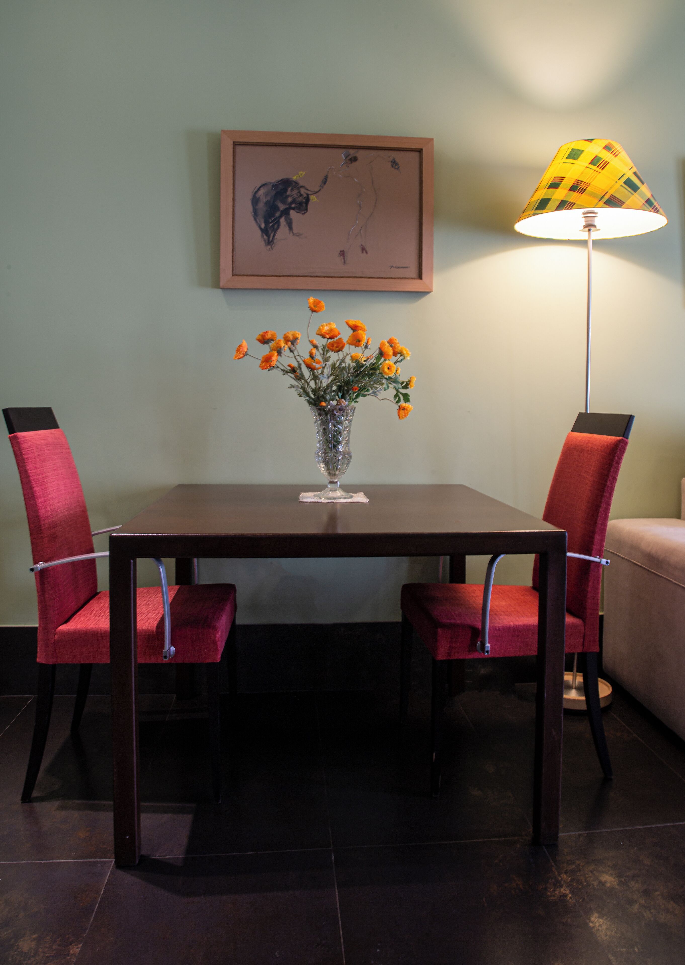 A dining table with two red chairs, a vase with orange flowers, a framed artwork on the wall, and a floor lamp.