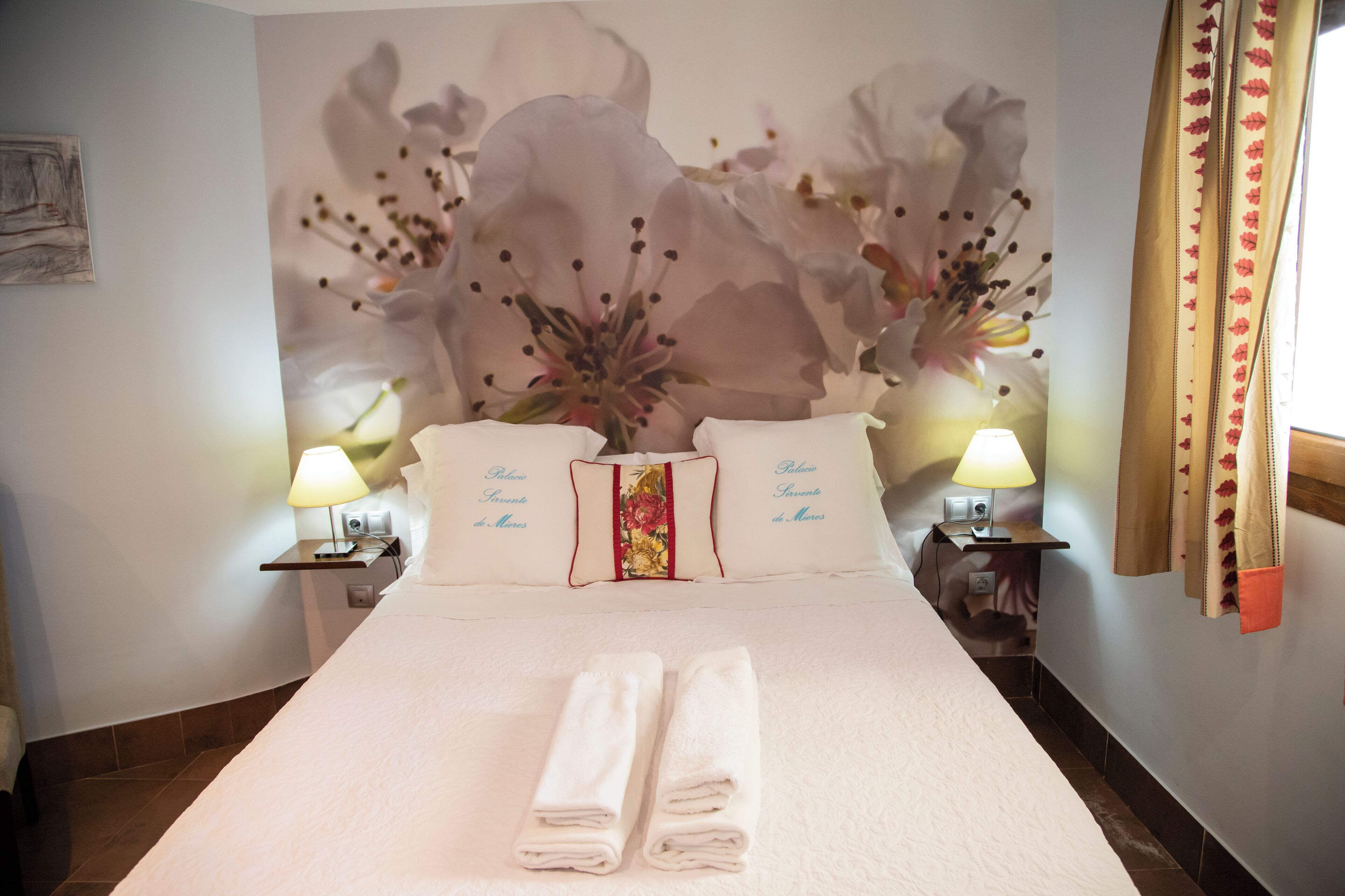 Una habitació d'hotel amb un llit gran, dues taules de nit amb làmpades i un mural floral a la paret.