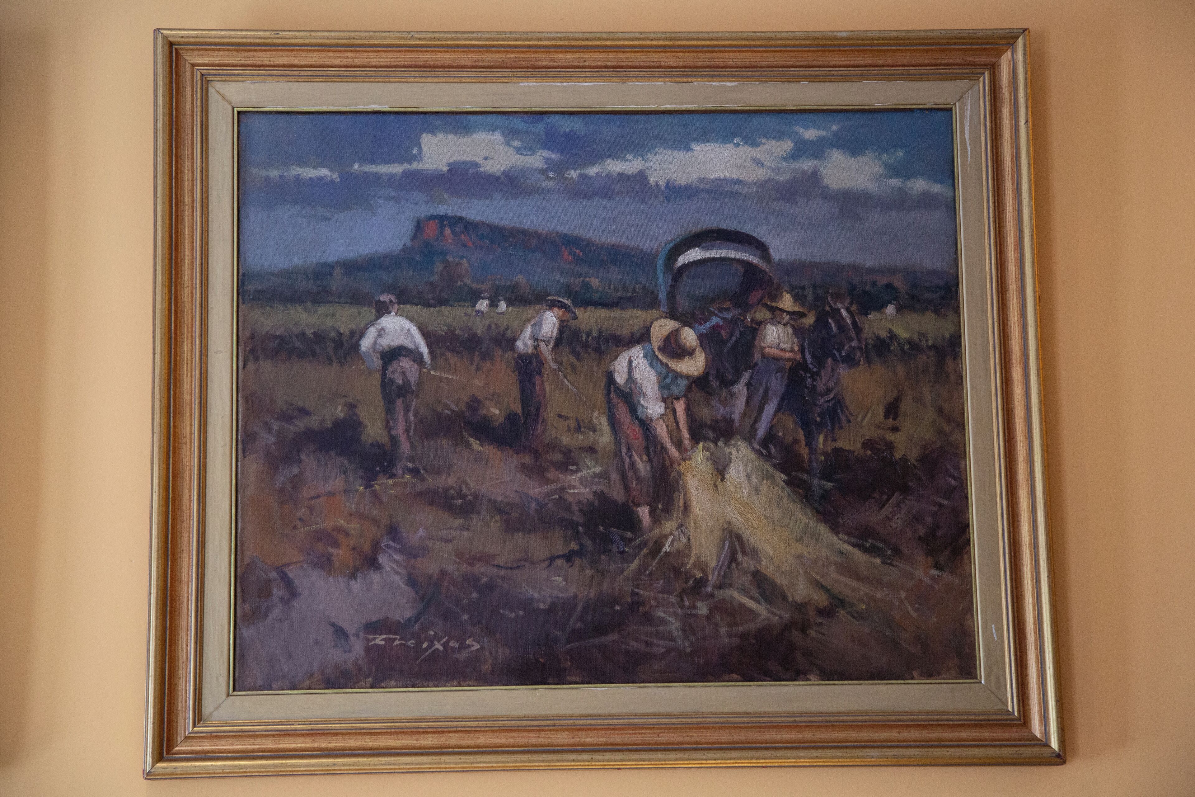 Ein gerahmtes Gemälde zeigt eine ländliche Szene mit arbeitenden Personen auf einem Feld, einem großen Eimer und einem Berg im Hintergrund.