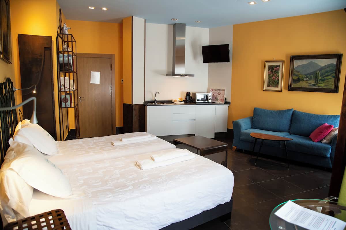 A hotel room with a bed, a sofa, a small kitchen area, and a TV mounted on the wall.