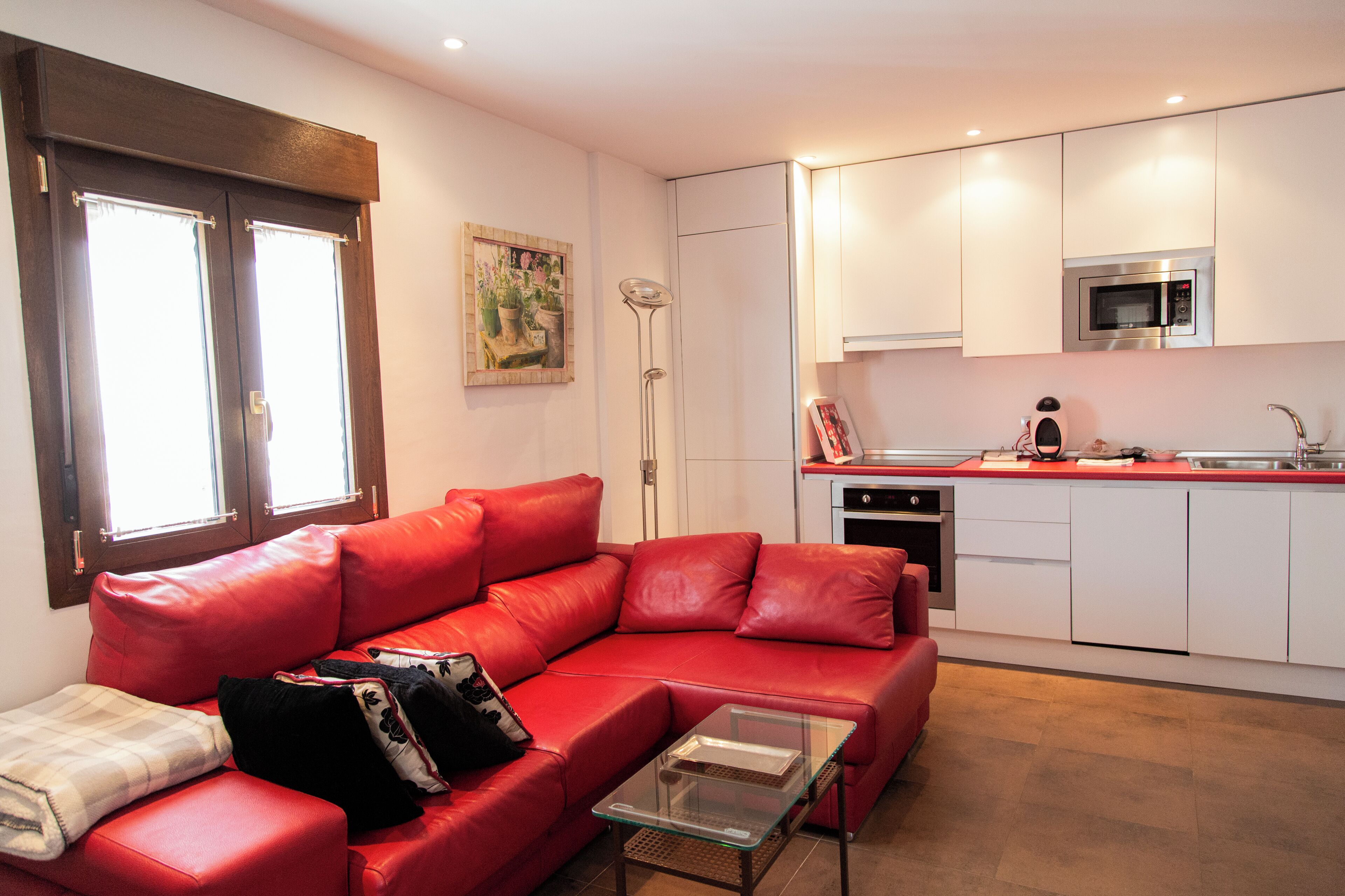 Ein modernes Wohnzimmer mit einem roten Ledersofa, einem Glastisch und einer Küchenzeile mit weißen Schränken und einer Edelstahl-Mikrowelle.