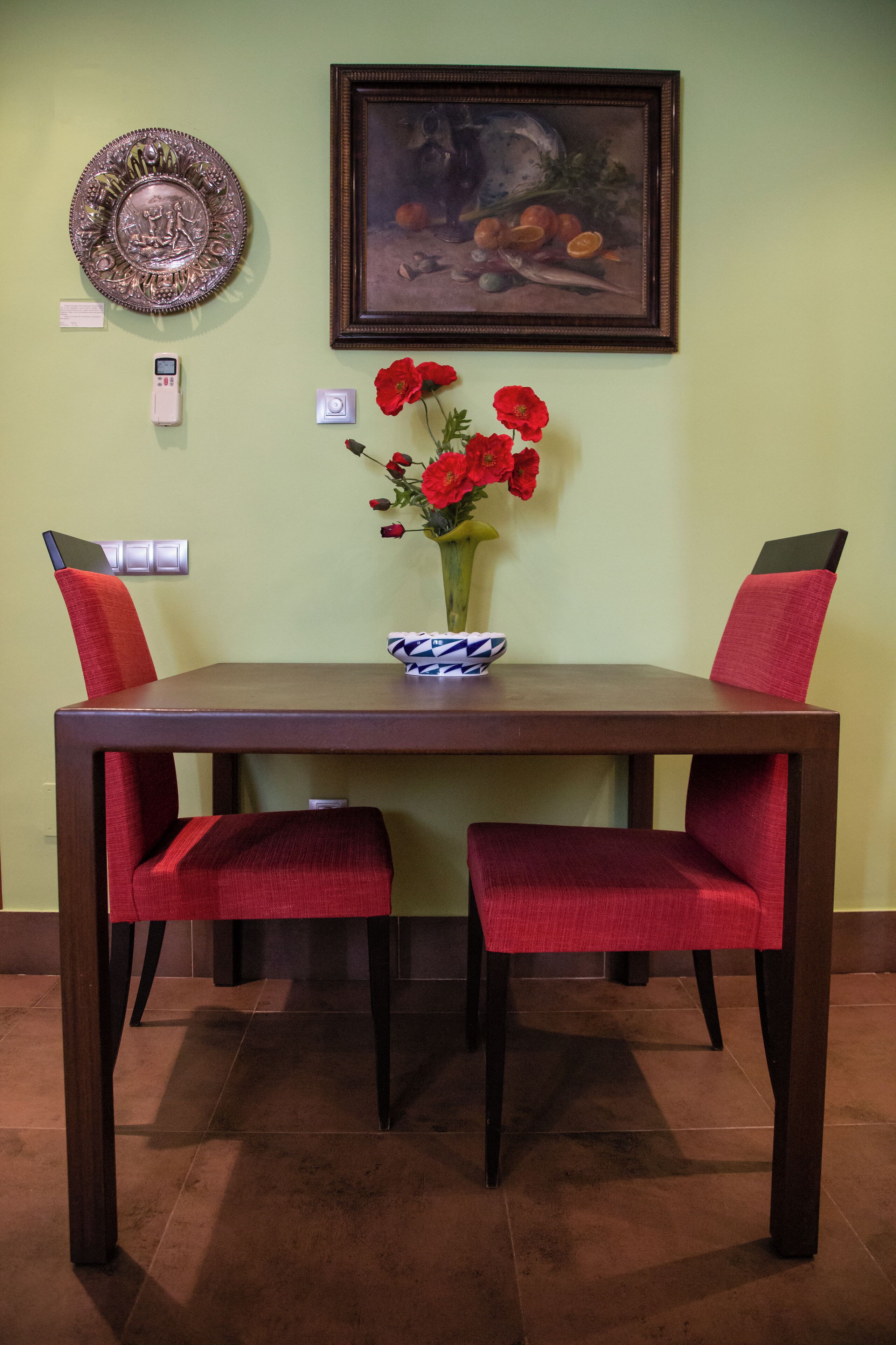 A dining table with two red chairs, a vase with red flowers, a framed painting, and a decorative plate on the wall.