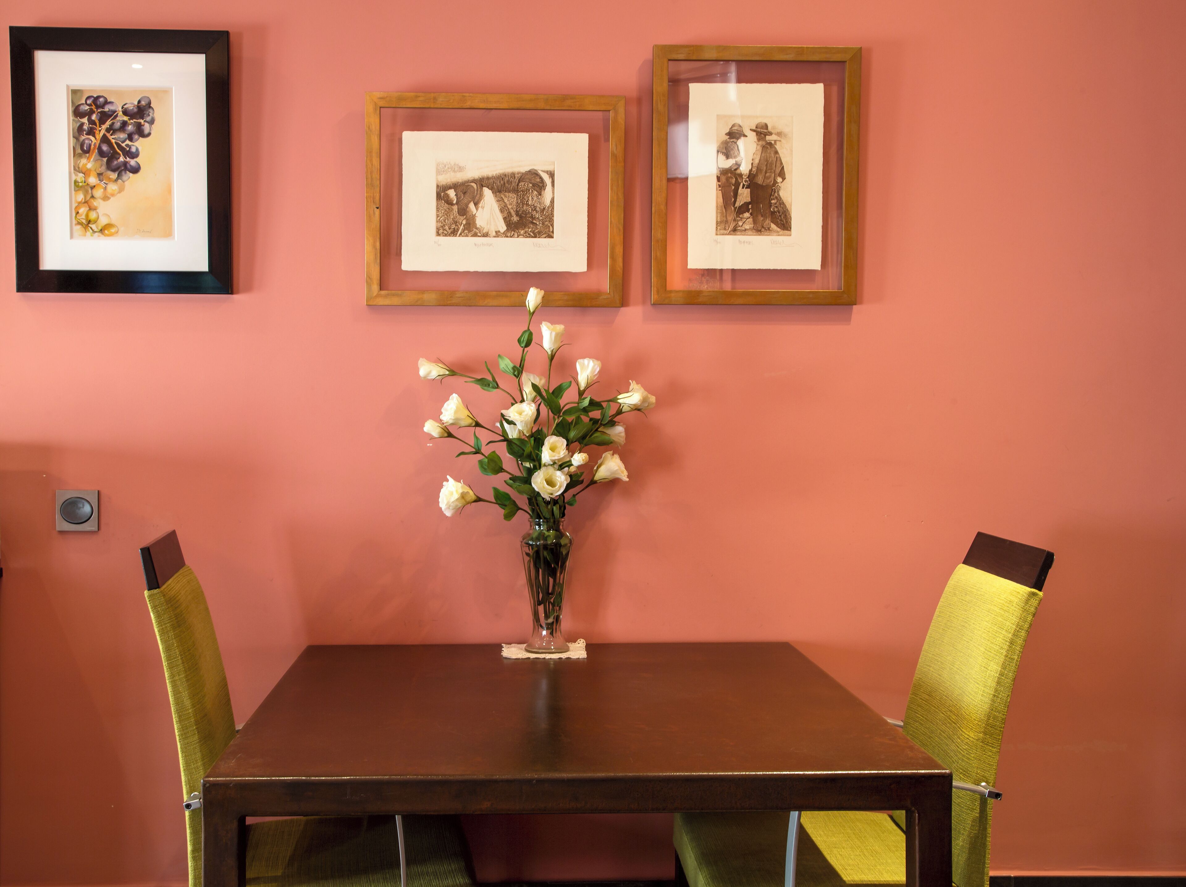 A dining area with a table, chairs, and a vase of flowers. There are framed pictures on the wall.