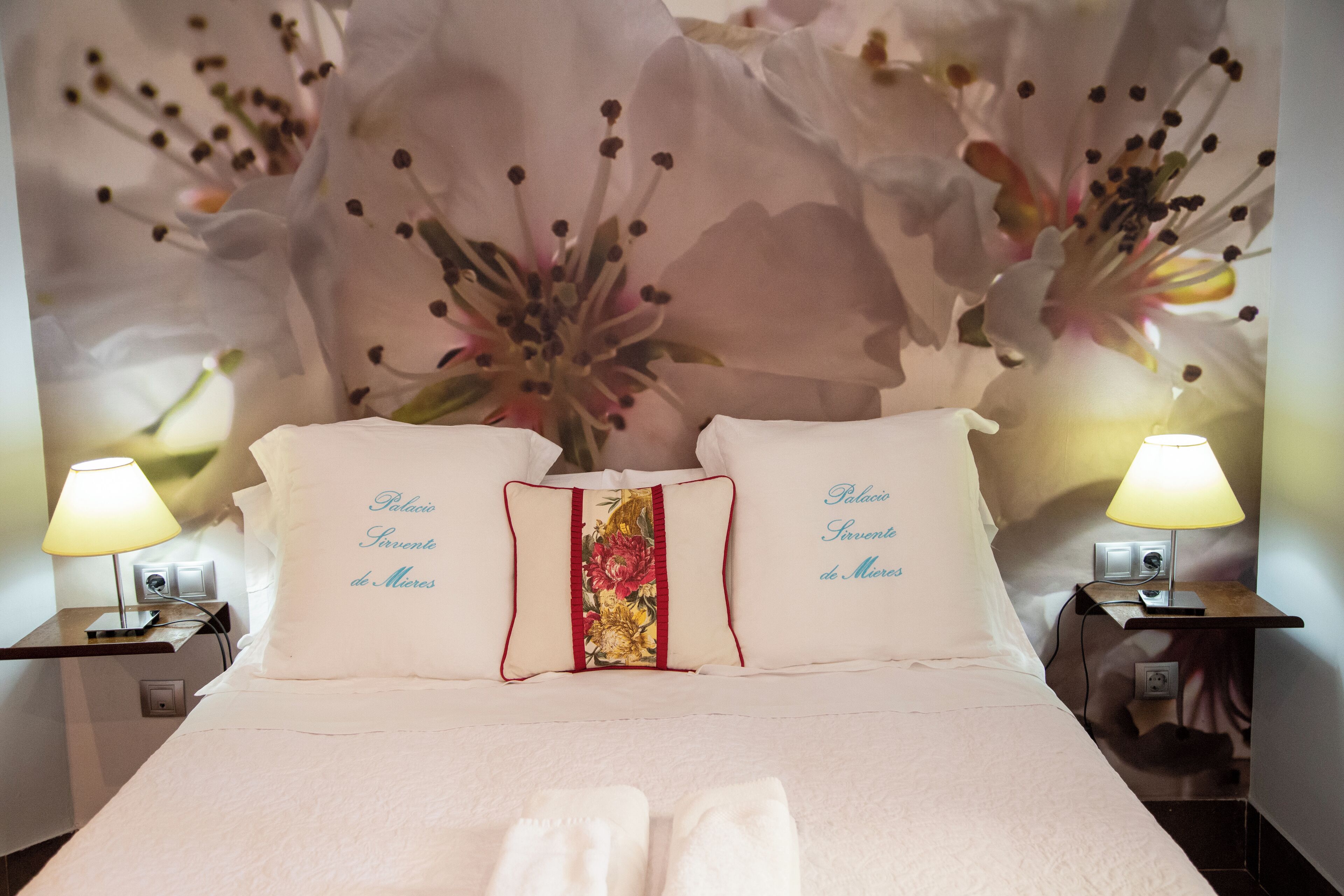 Une chambre d’hôtel avec un lit, deux tables de chevet, des lampes et une décoration murale florale.