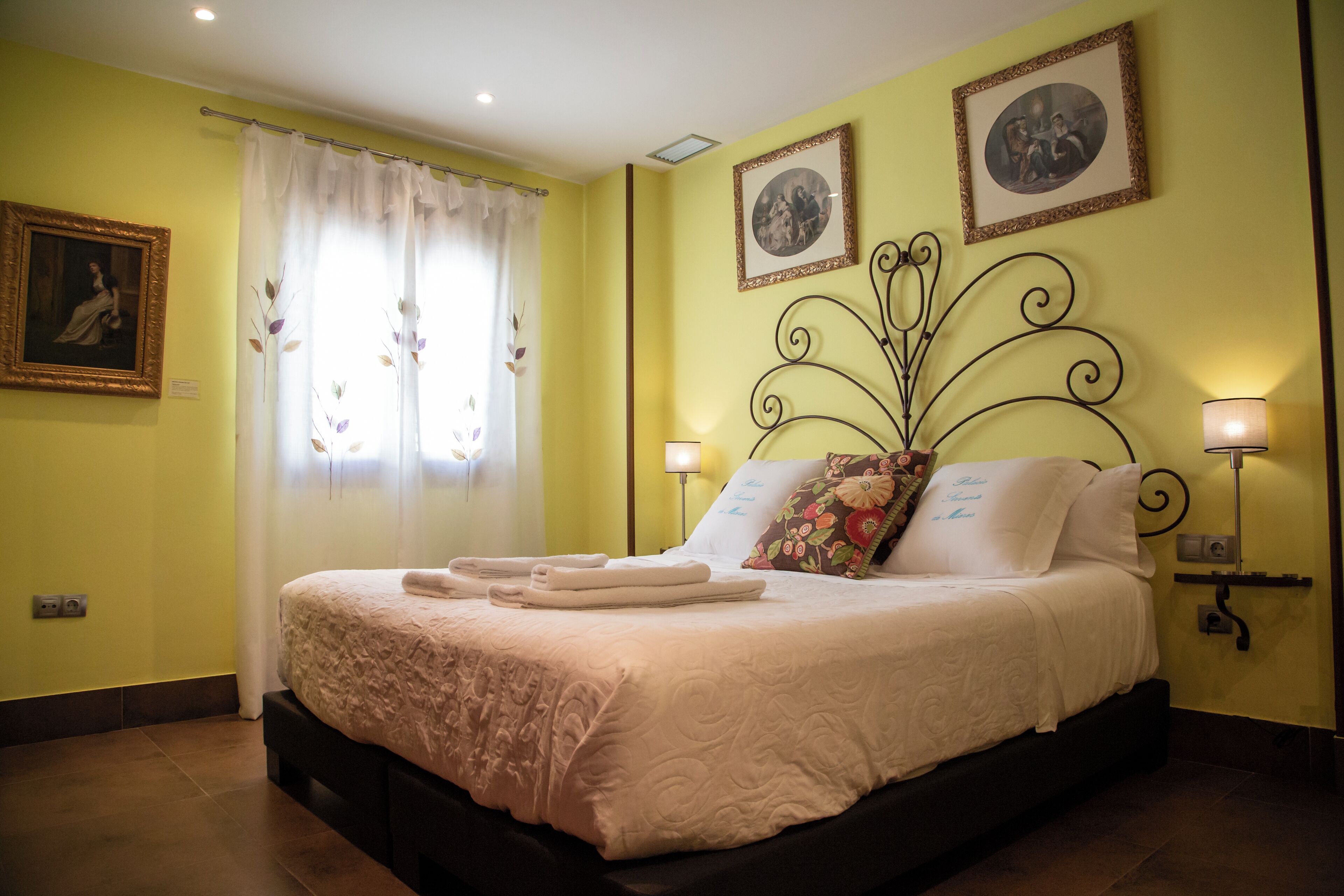 Una cama bien tendida con un cojín floral, una mesita de noche con una lámpara y dos cuadros enmarcados en la pared.