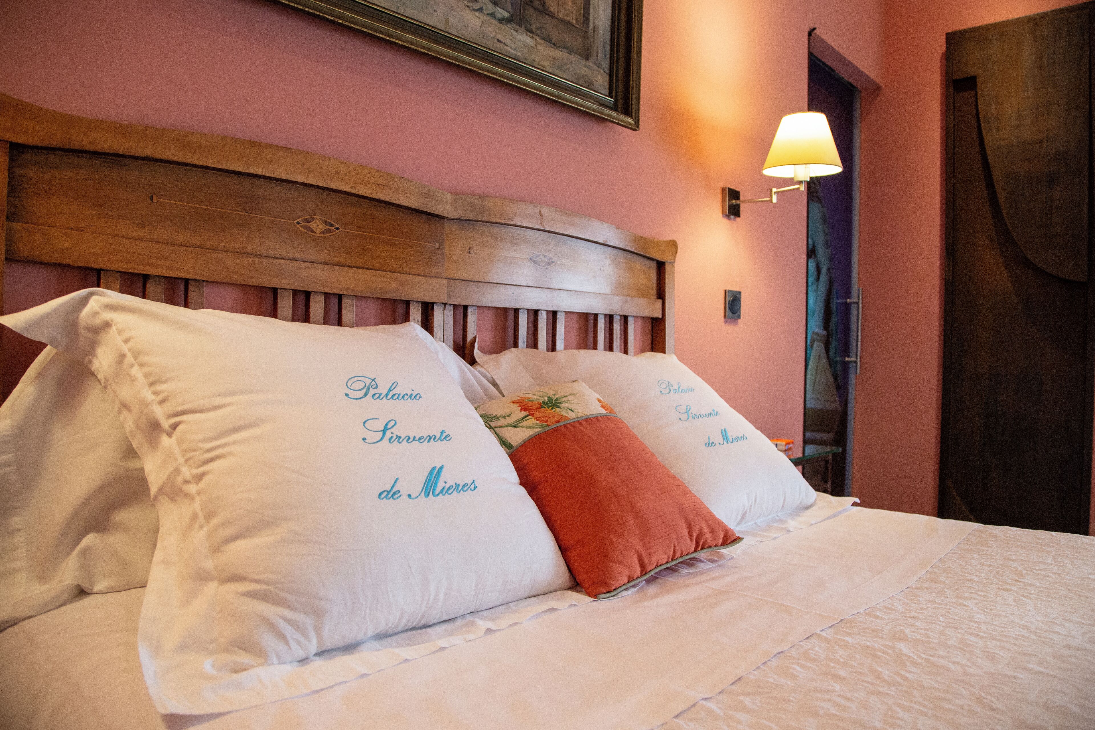 Una cama con almohadas blancas y naranjas, un cabecero de madera y una lámpara fijada a la pared.