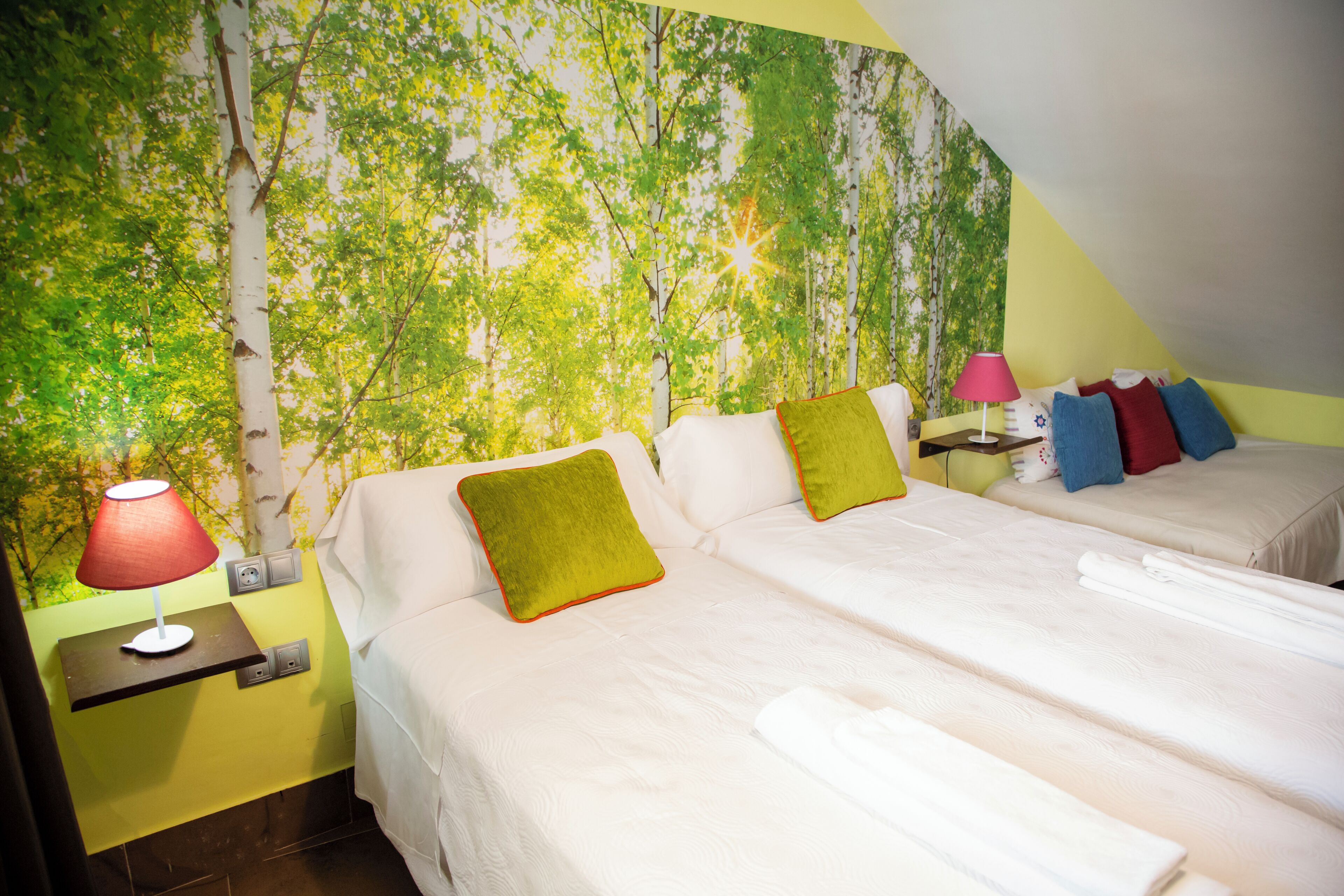 A room with two beds, a nightstand with a lamp, and a wall mural of trees.