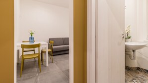 Une pièce comprenant un coin repas avec une table blanche et deux chaises, un canapé gris et un vase avec des fleurs posé sur la table.