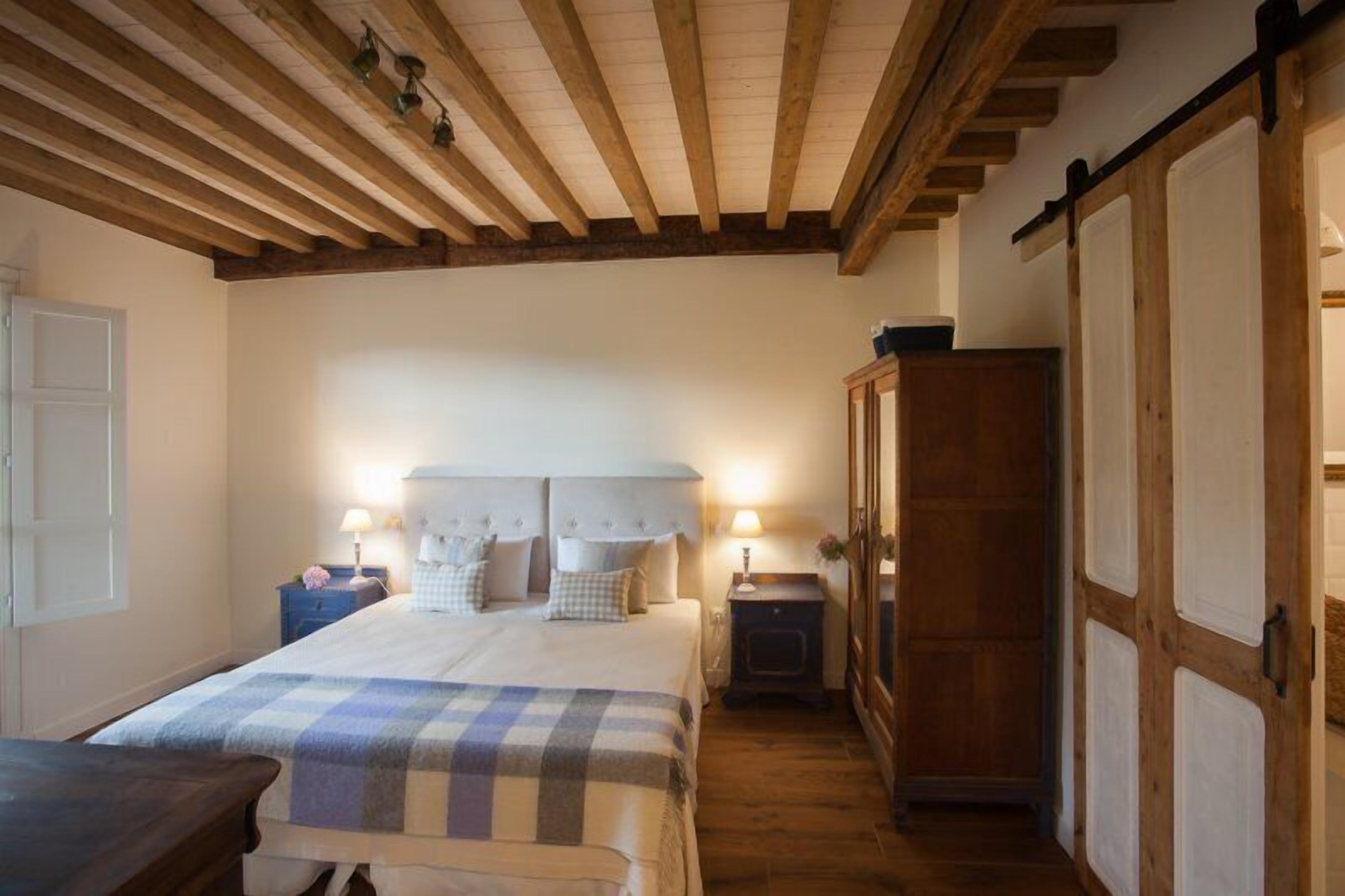 Une chambre avec un grand lit, des poutres apparentes en bois au plafond, une armoire et des tables de chevet avec des lampes.