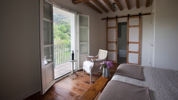 A room with a wooden floor, a bed, a chair, a small table with a vase, and a sliding door leading to a balcony with a view of greenery. - Posada Seis Leguas (Cartes)