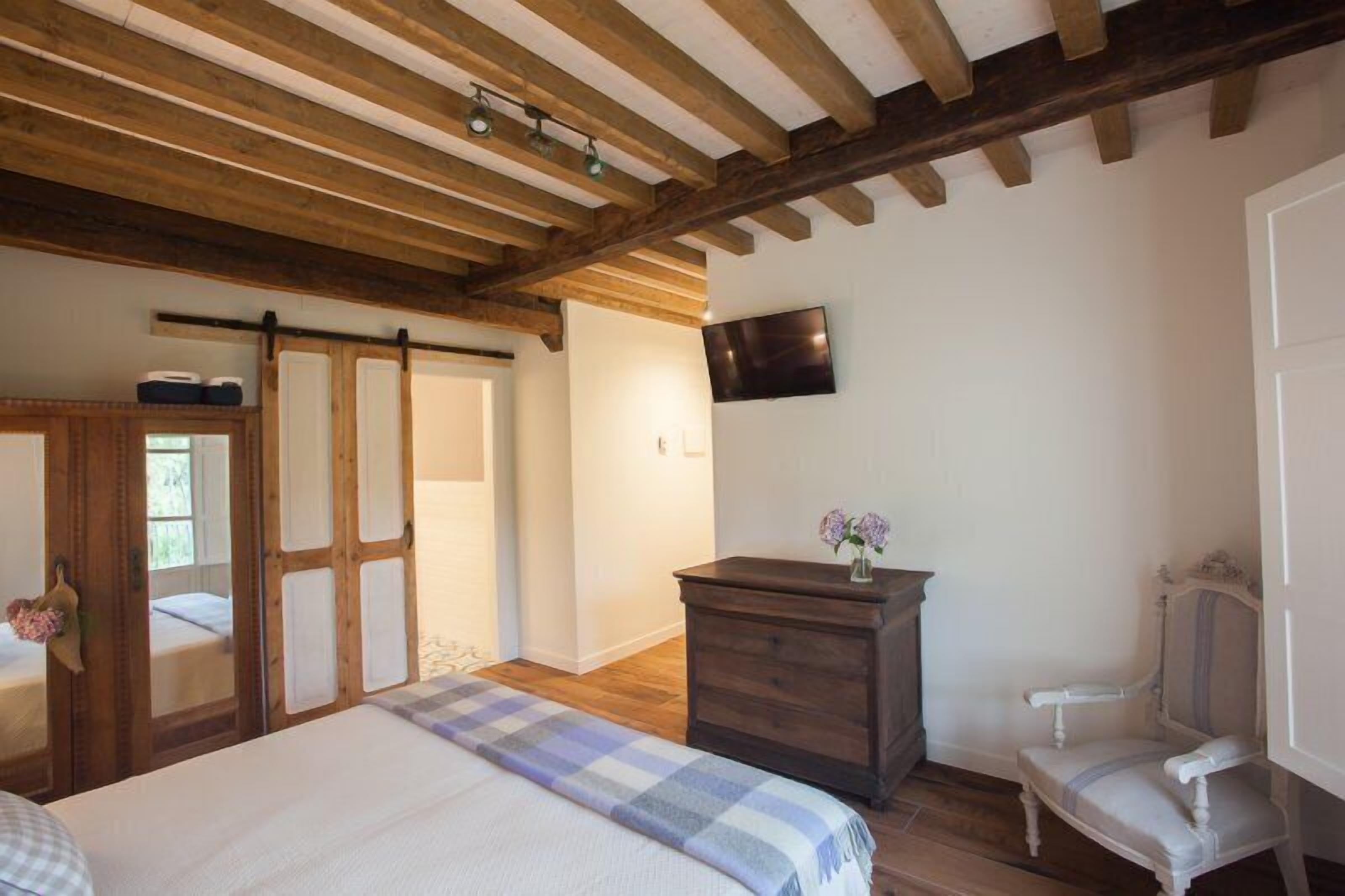 A bedroom with a bed, wooden ceiling beams, a dresser, a chair, and a television on the wall.