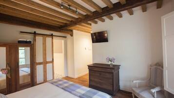 A bedroom with a bed, wooden ceiling beams, a dresser, a chair, and a television on the wall.