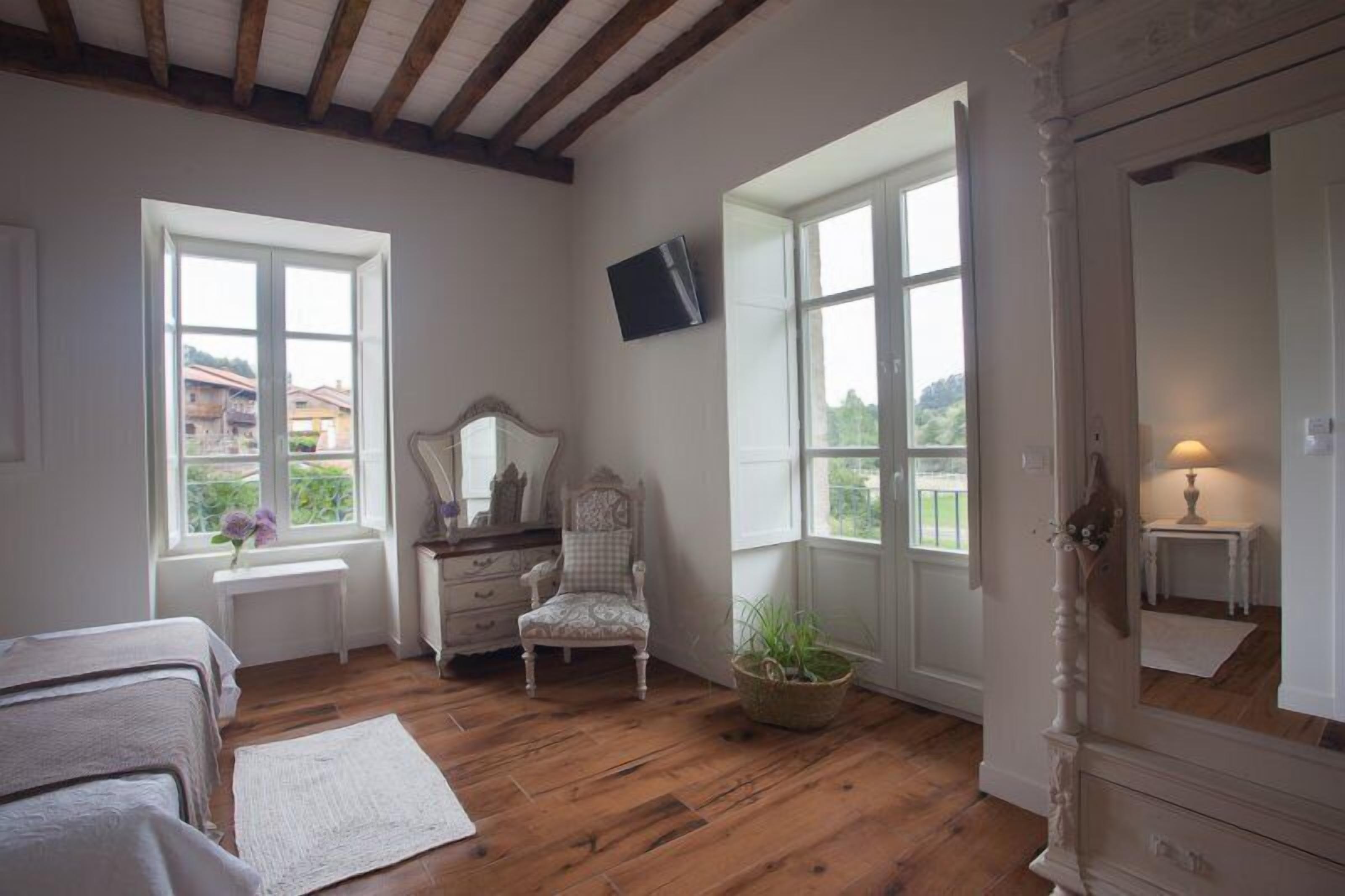 A bedroom with wooden flooring, a bed, a chair, a dresser, a television, and large windows with a view of the outside.