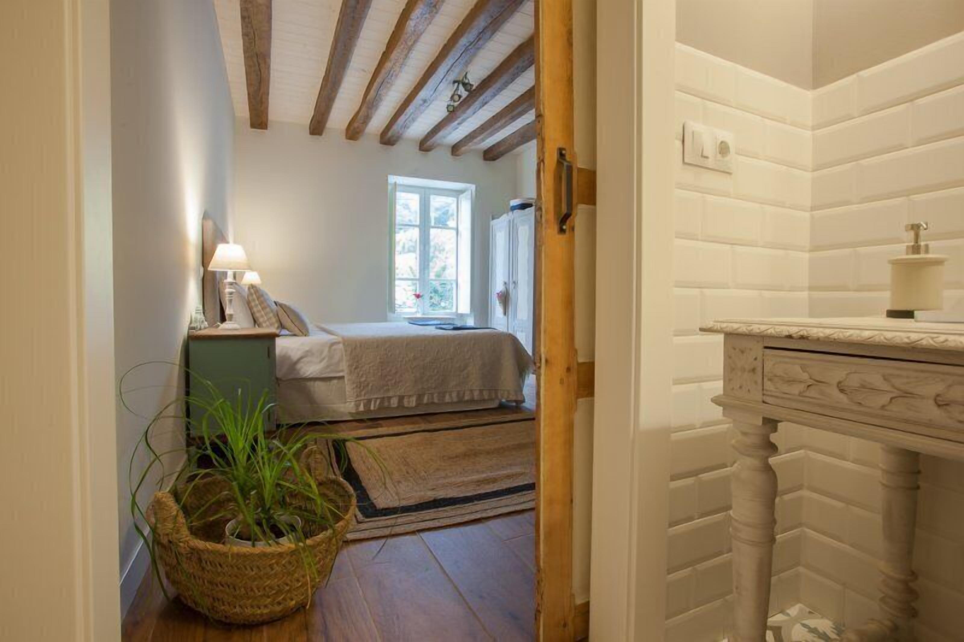 A narrow hallway leading to a bedroom with a window, a bedside table with a lamp, and a wicker basket on the floor.
