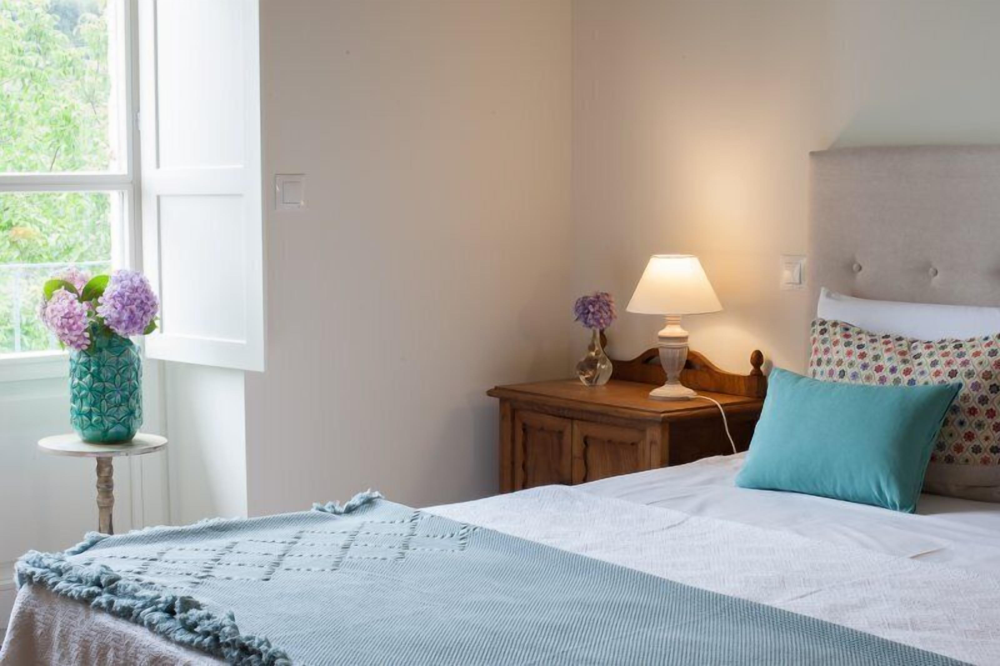 A bedroom with a bed, a bedside table with a lamp, a vase with flowers, and a window with a view of greenery.