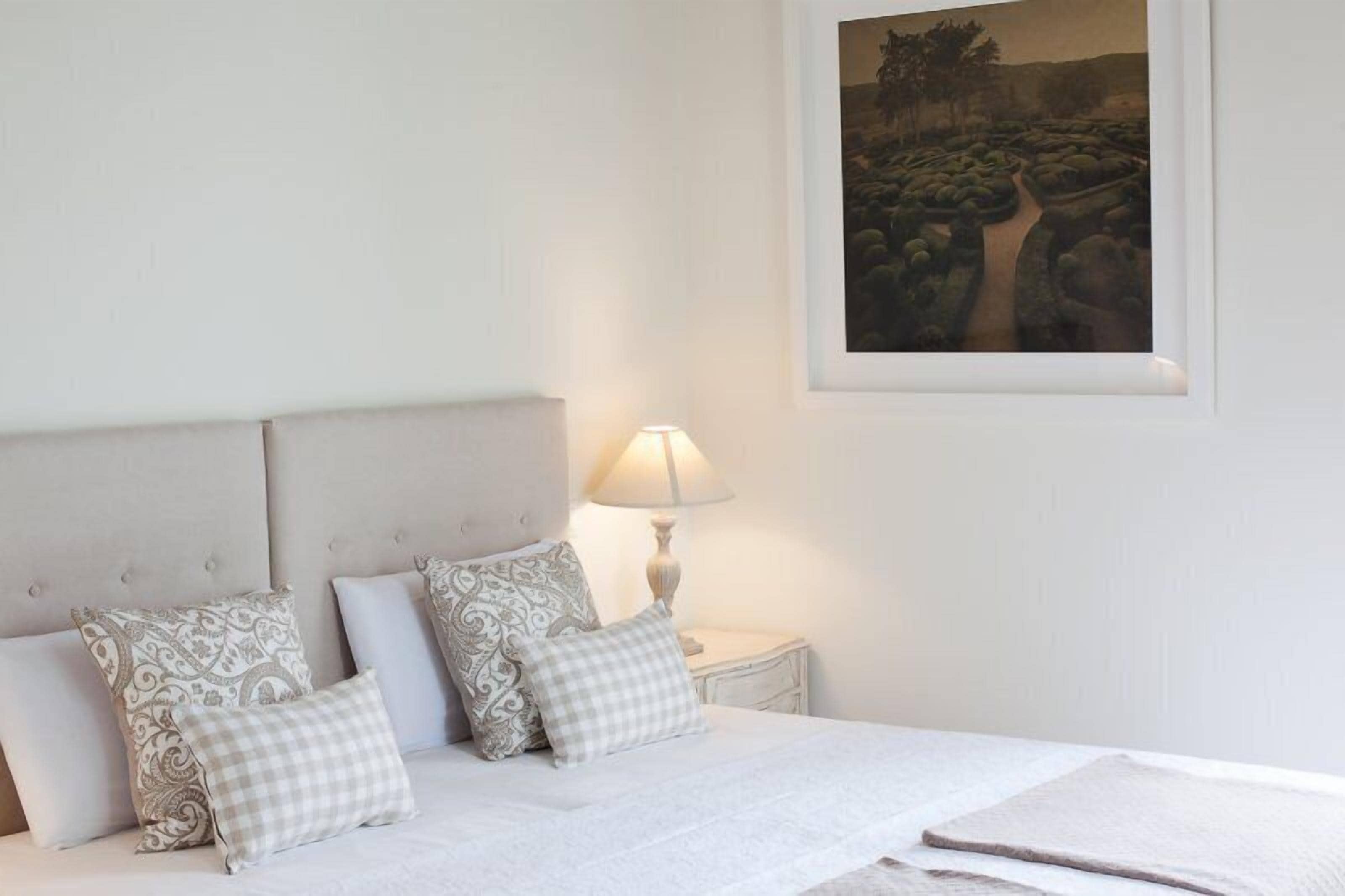 Ein ordentlich bezogenes Bett mit gemusterten Kissen, einer Nachttischlampe und einem gerahmten Landschaftsbild an der Wand.