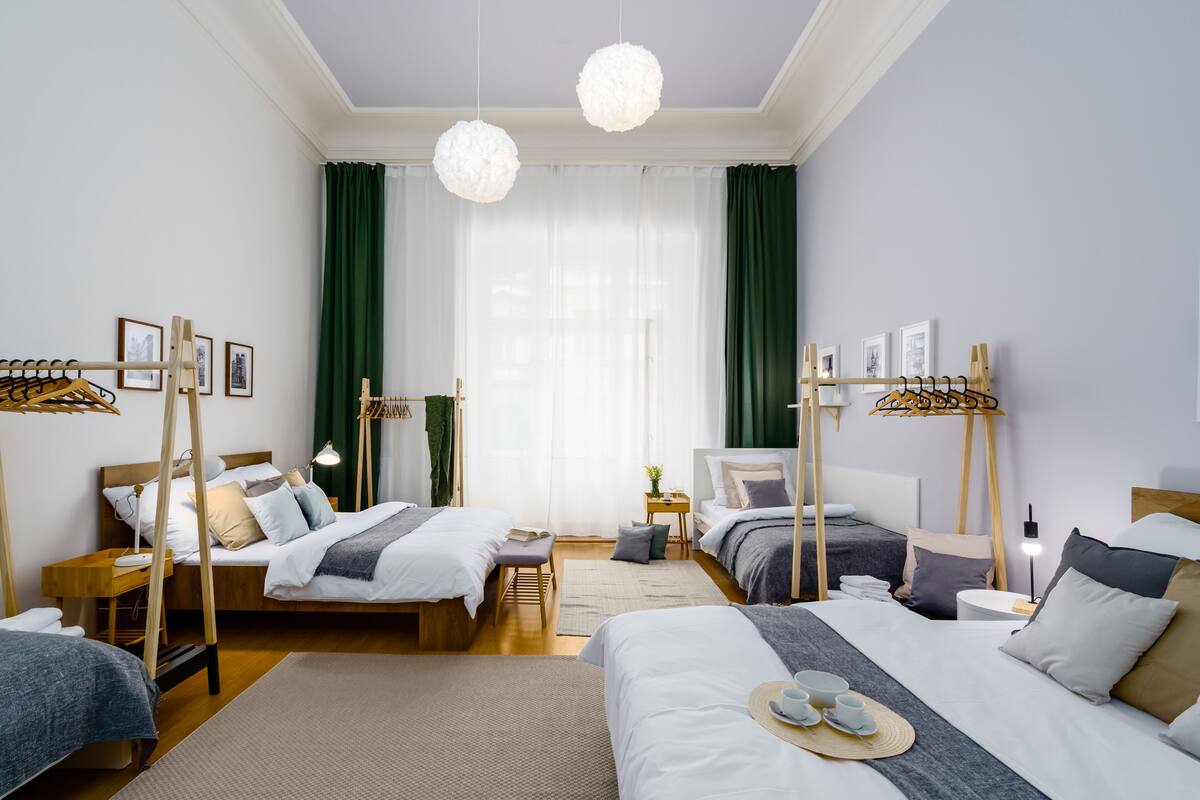 A hotel room with two beds, a wooden headboard, a bedside table with a lamp, a small table with a plant, and a large window with curtains.