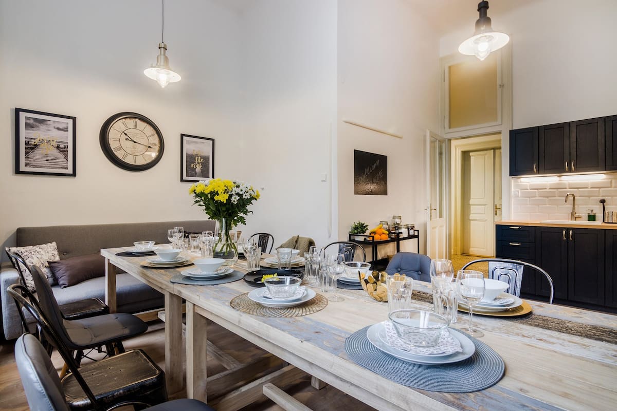 A dining area with a long table set for a meal, a sofa, and a kitchen in the background.