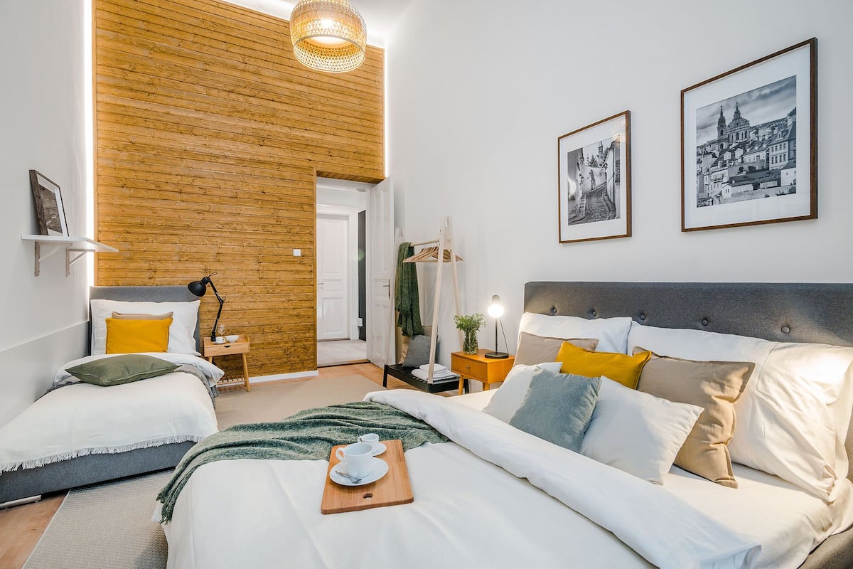 A modern hotel room with a wooden accent wall, a large bed with multiple pillows, a bedside table with a lamp, and two framed pictures on the wall.