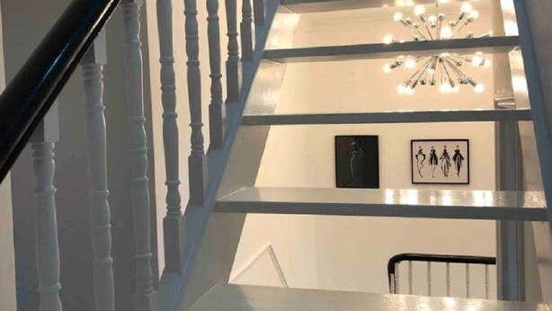Una escalera con balaustradas blancas y una barandilla negra que lleva a un rellano con un cuadro enmarco y un pequeño banco.