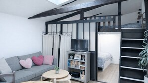 Ein Loft-Zimmer mit hohen Decken, freiliegenden Balken, einem Oberlicht, einer Couch mit Kissen, einem kleinen Tisch, einem Bücherregal, einem Fernseher und einer Treppe.