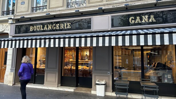 Ein Schaufenster mit einem schwarz-weiß gestreiften Vordach, auf dem die Namen 'BOULANGERIE' und 'GANA' zu sehen sind, und eine Frau steht davor.