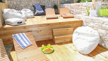 Une terrasse en bois avec un muret en pierre, un banc en bois, un pouf et une petite table avec des boissons.
