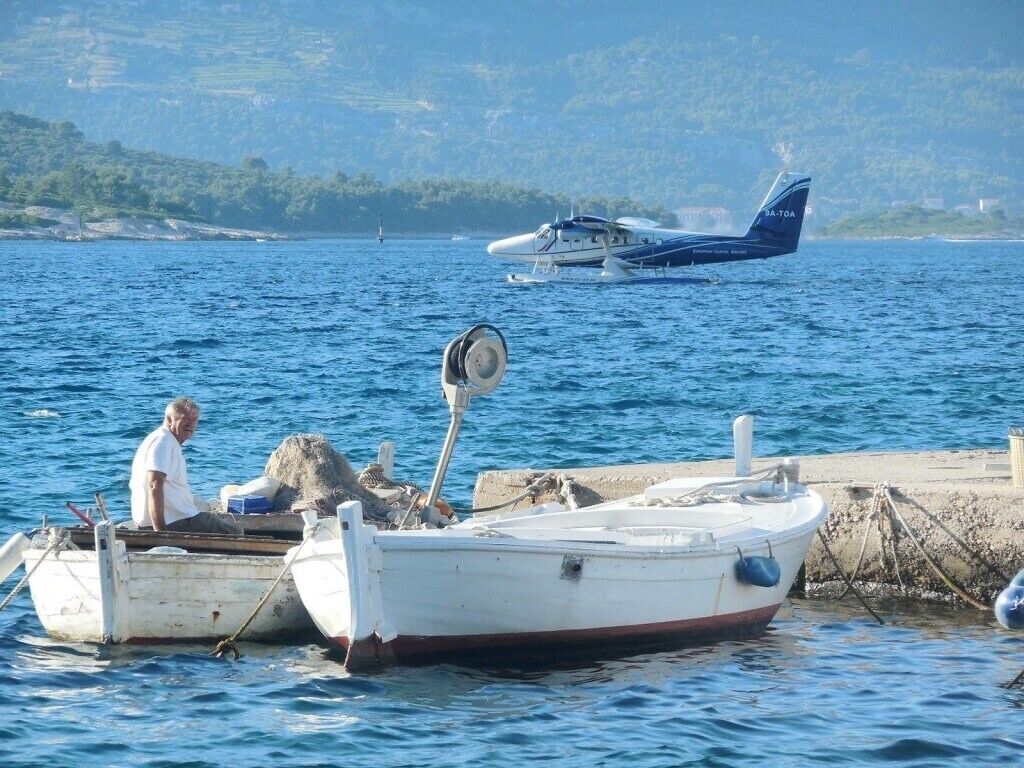 Un petit bateau était amarré à un quai, avec une personne à bord. On pouvait apercevoir un hydravion en arrière-plan et, au loin, un paysage vallonné.