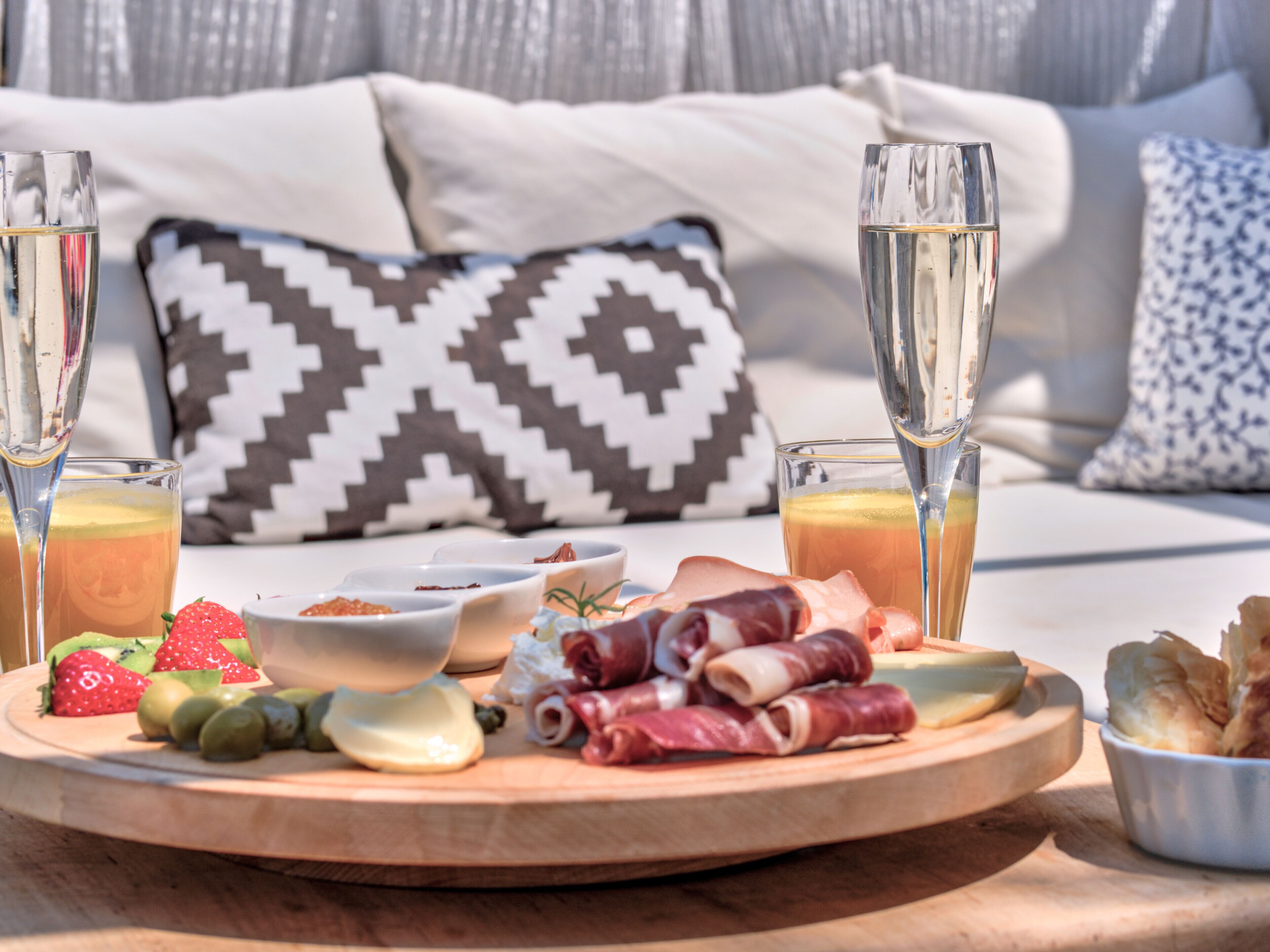 Une table dressée pour le petit-déjeuner, avec une planche en bois sur laquelle sont disposés des fruits, du fromage et de la charcuterie, accompagnée de deux verres de jus d’orange et de deux coupes à champagne.