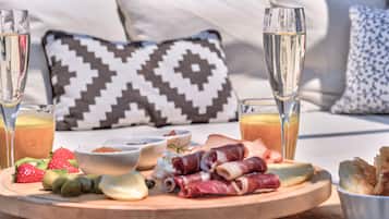 Une table dressée pour le petit-déjeuner, avec une planche en bois sur laquelle sont disposés des fruits, du fromage et de la charcuterie, accompagnée de deux verres de jus d’orange et de deux coupes à champagne.