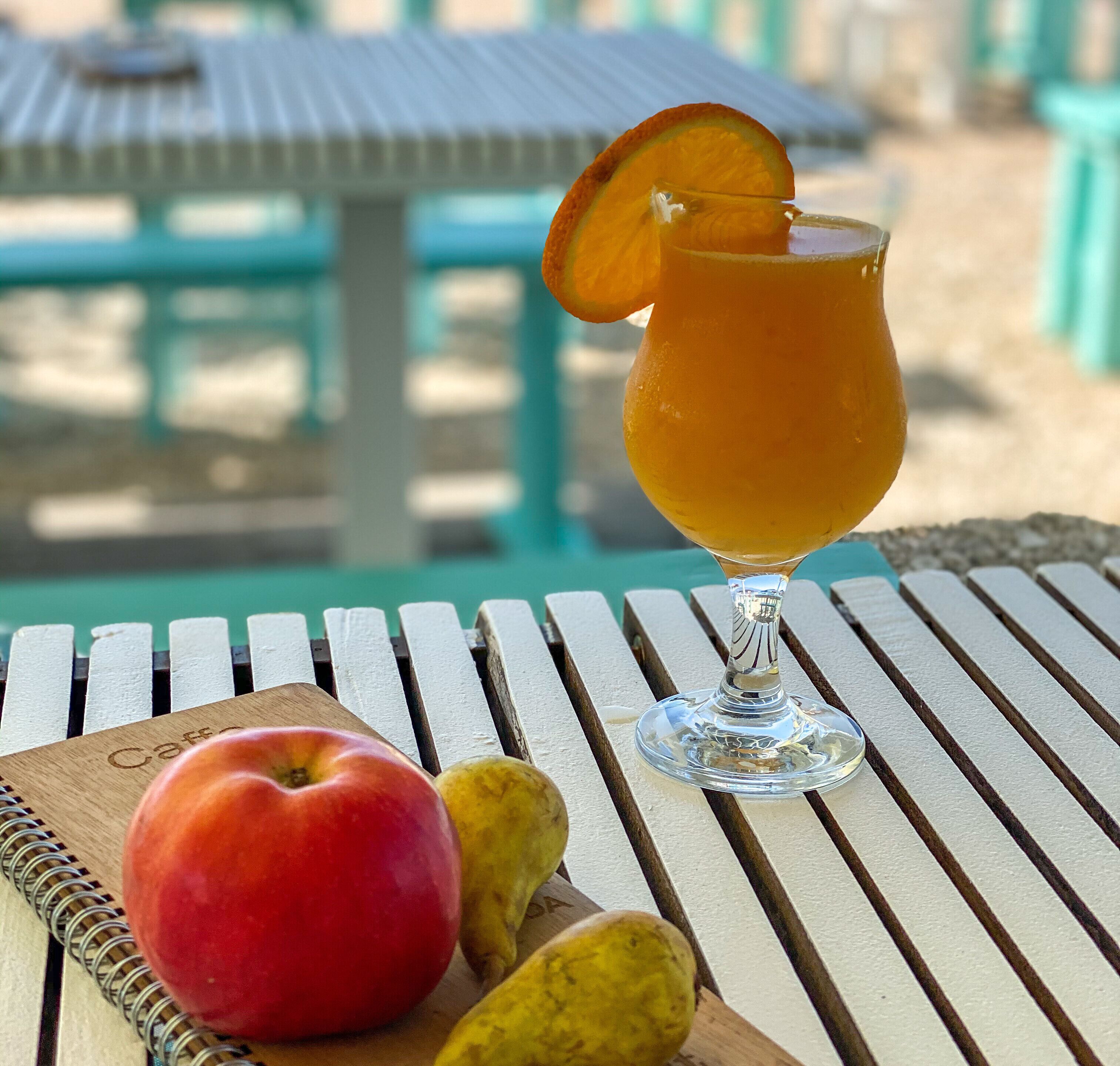A table with a glass of orange juice, a red apple, and a yellow fruit on a wooden board.