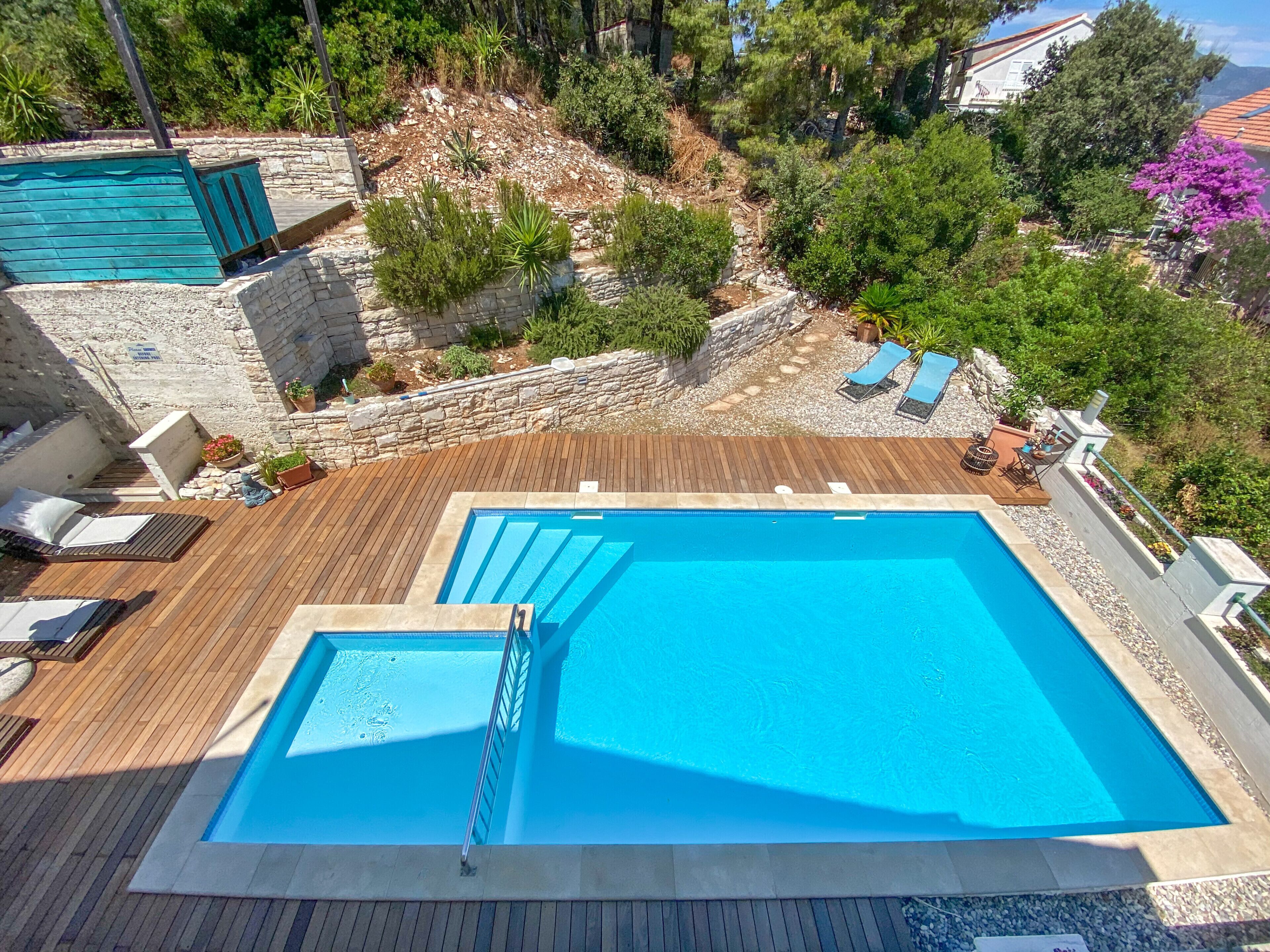 Ein rechteckiger Pool mit Holzdeck und Liegestühlen, umgeben von Steinmauern und Grünpflanzen.