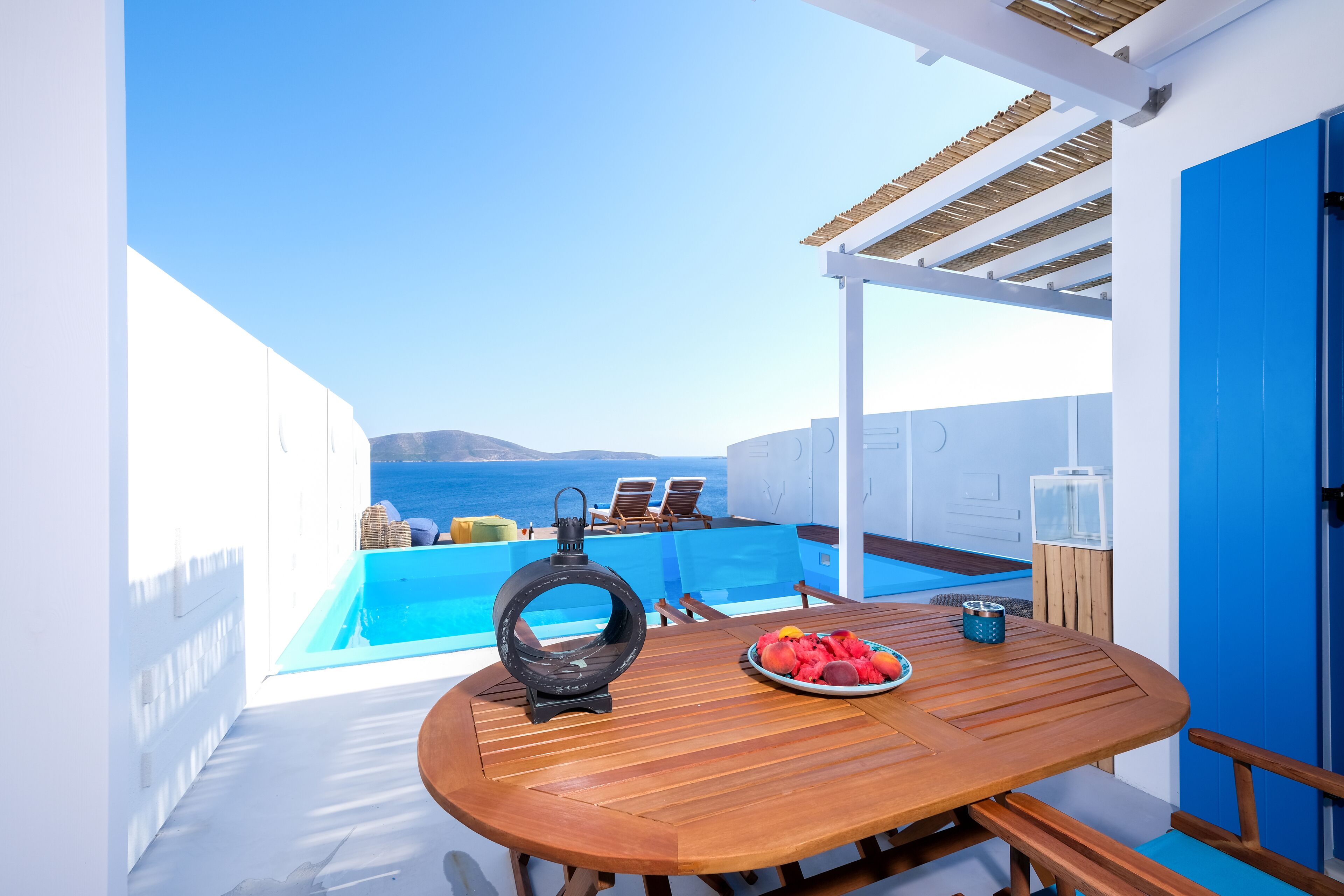 A wooden table with a plate of fruit and a lantern on a balcony overlooking a pool and the sea.