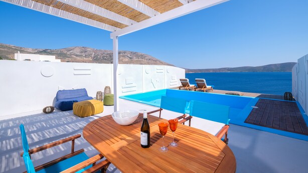 A sunlit outdoor seating area with a wooden table, chairs, and a view of the sea.