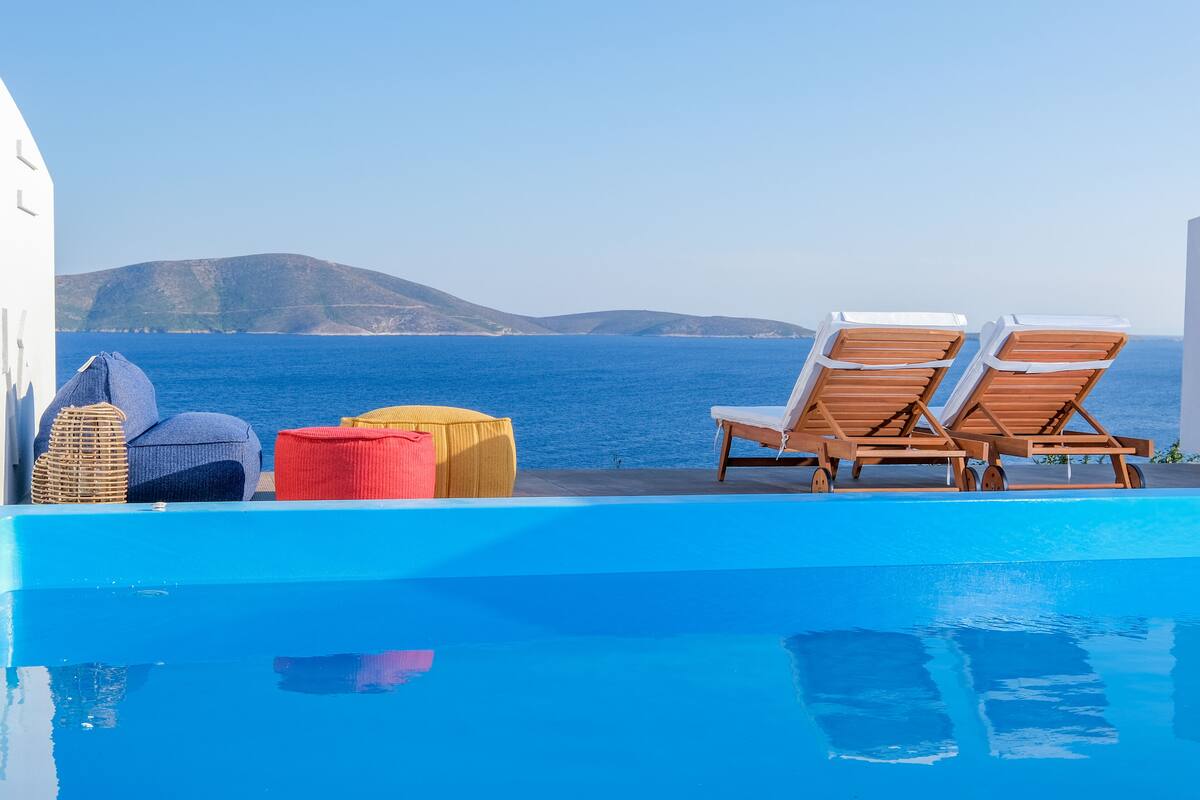 Un espace piscine doté de chaises longues et de poufs, offrant une vue sur un plan d’eau et des collines.