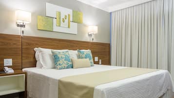 A neatly made bed with a white and beige bedspread, two pillows, and a wooden headboard. There is a bedside table with a lamp and a phone. The room has wooden flooring and a large window with curtains.