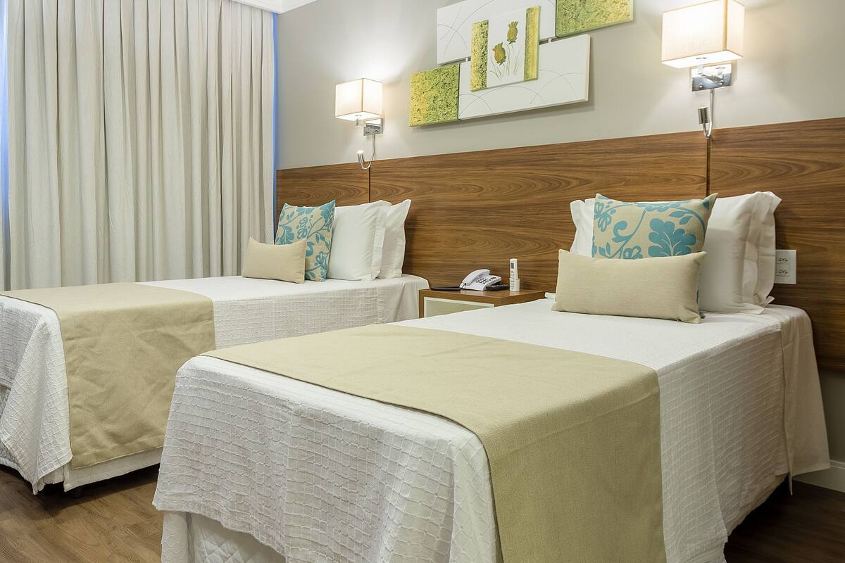 Une chambre d’hôtel avec deux lits, un plancher en bois, une tête de lit en bois, une lampe fixée au mur et un rideau.