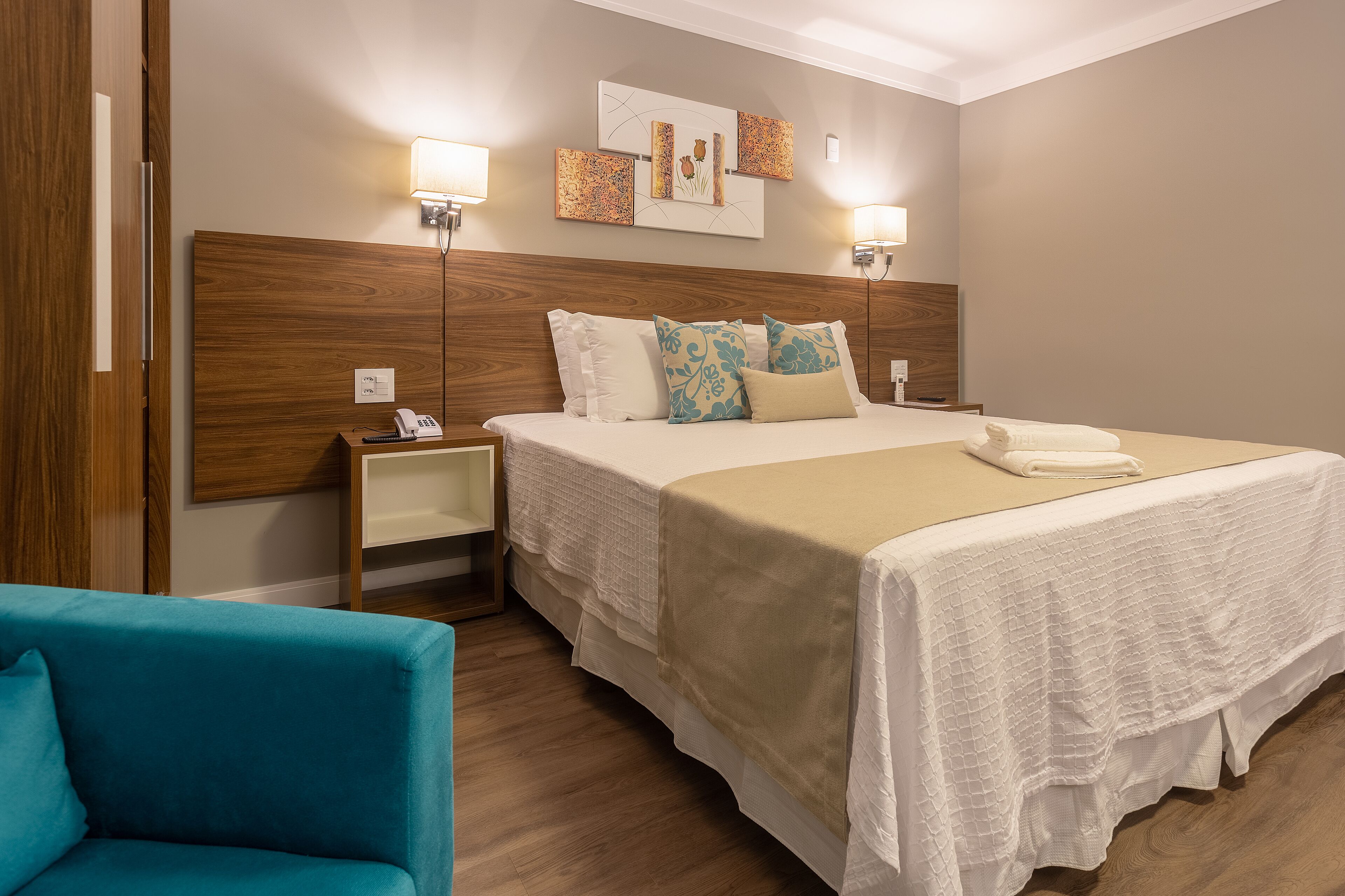 Quarto de hotel com uma cama grande, duas mesas de cabeceira, um criado-mudo, uma poltrona azul e uma obra de arte pendurada na parede.