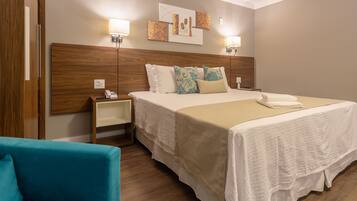 Quarto de hotel com uma cama grande, duas mesas de cabeceira, um criado-mudo, uma poltrona azul e uma obra de arte pendurada na parede.