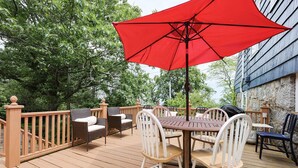 Eine Holzterrasse mit Gartenmöbeln, darunter Stühle und ein Tisch, und ein roter Sonnenschirm, der Schatten spendet.