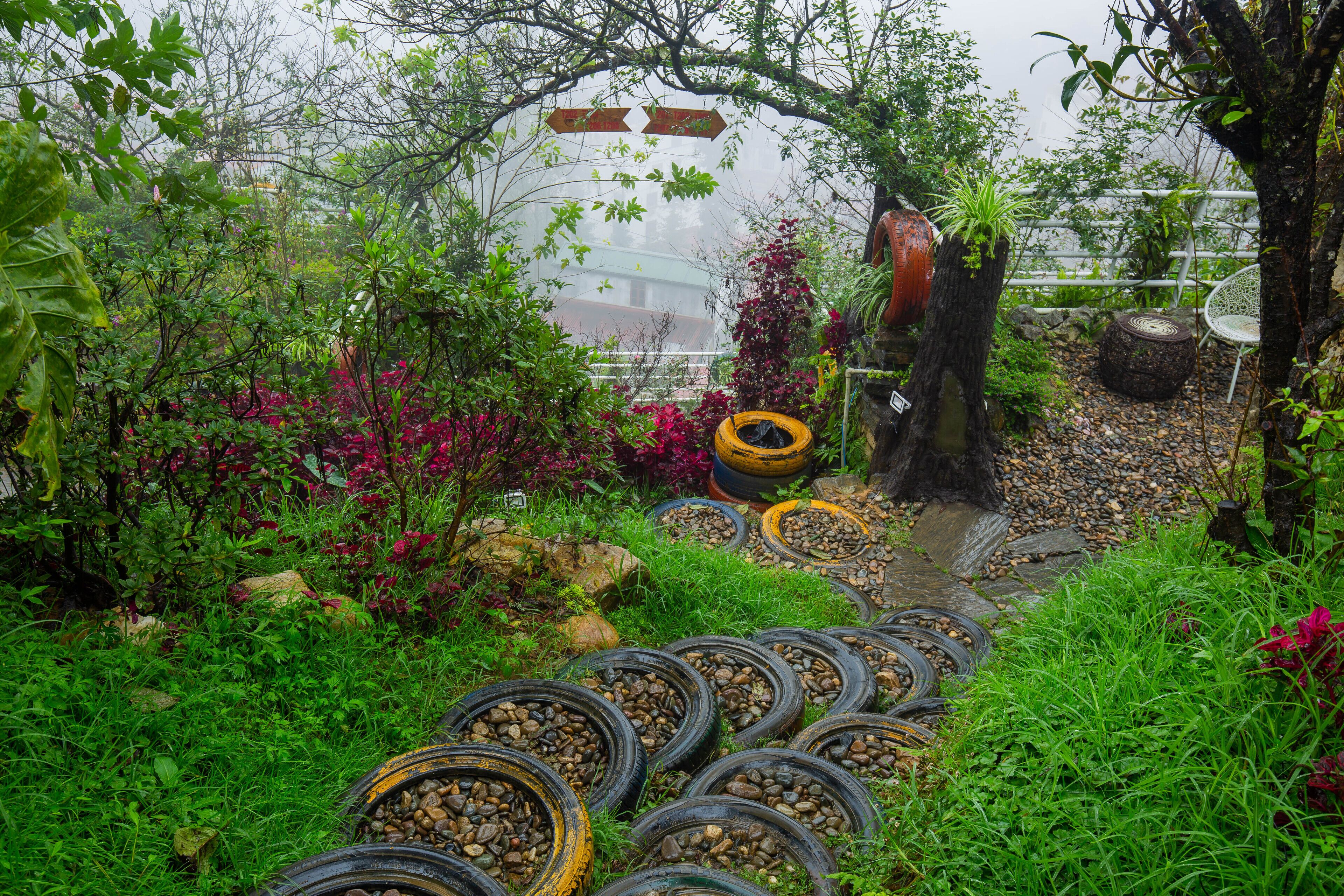 A garden pathway lined with tires filled with pebbles, surrounded by lush greenery and flowering plants.