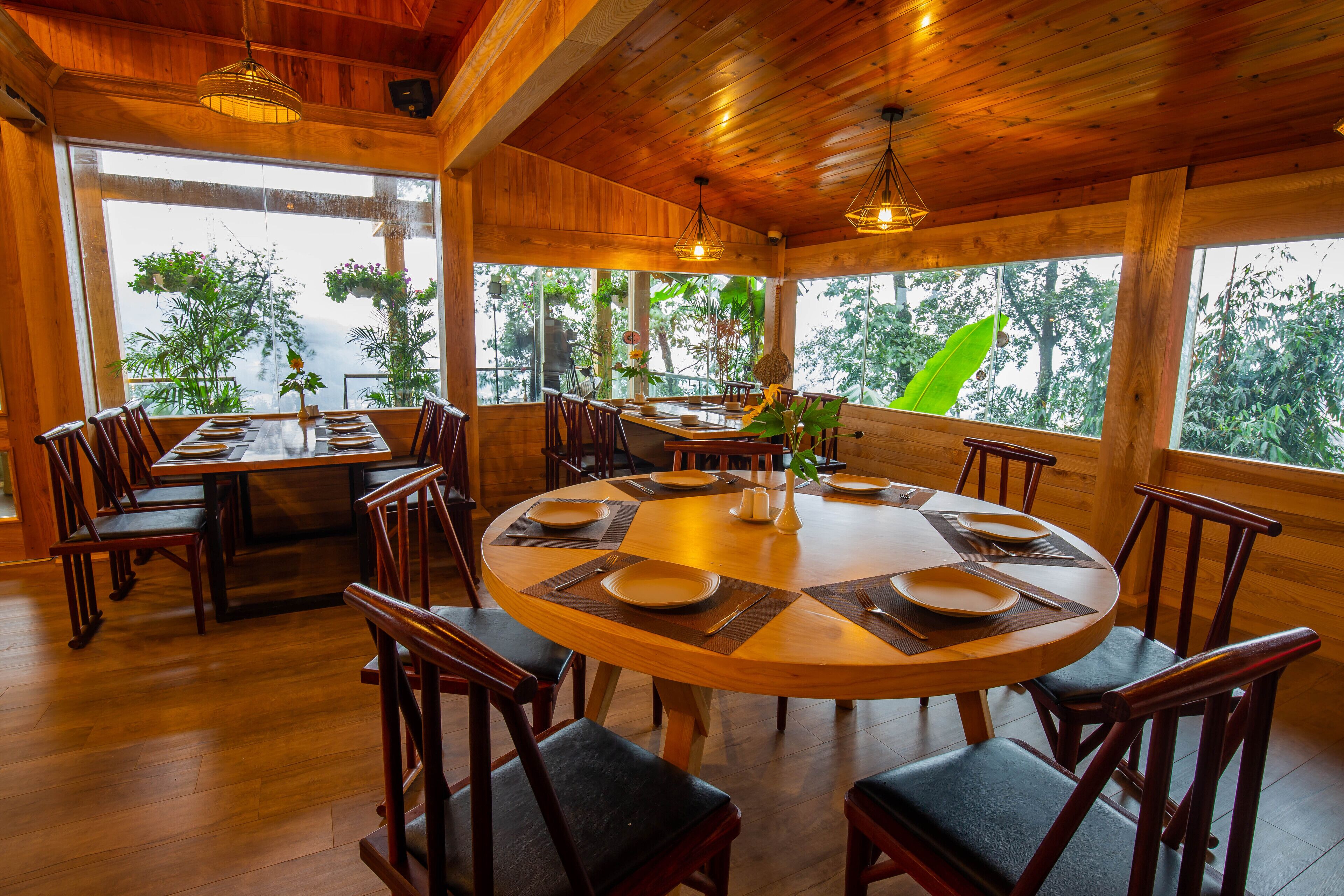 A wooden dining area with round tables and chairs, large windows with views of greenery, and hanging pendant lights.
