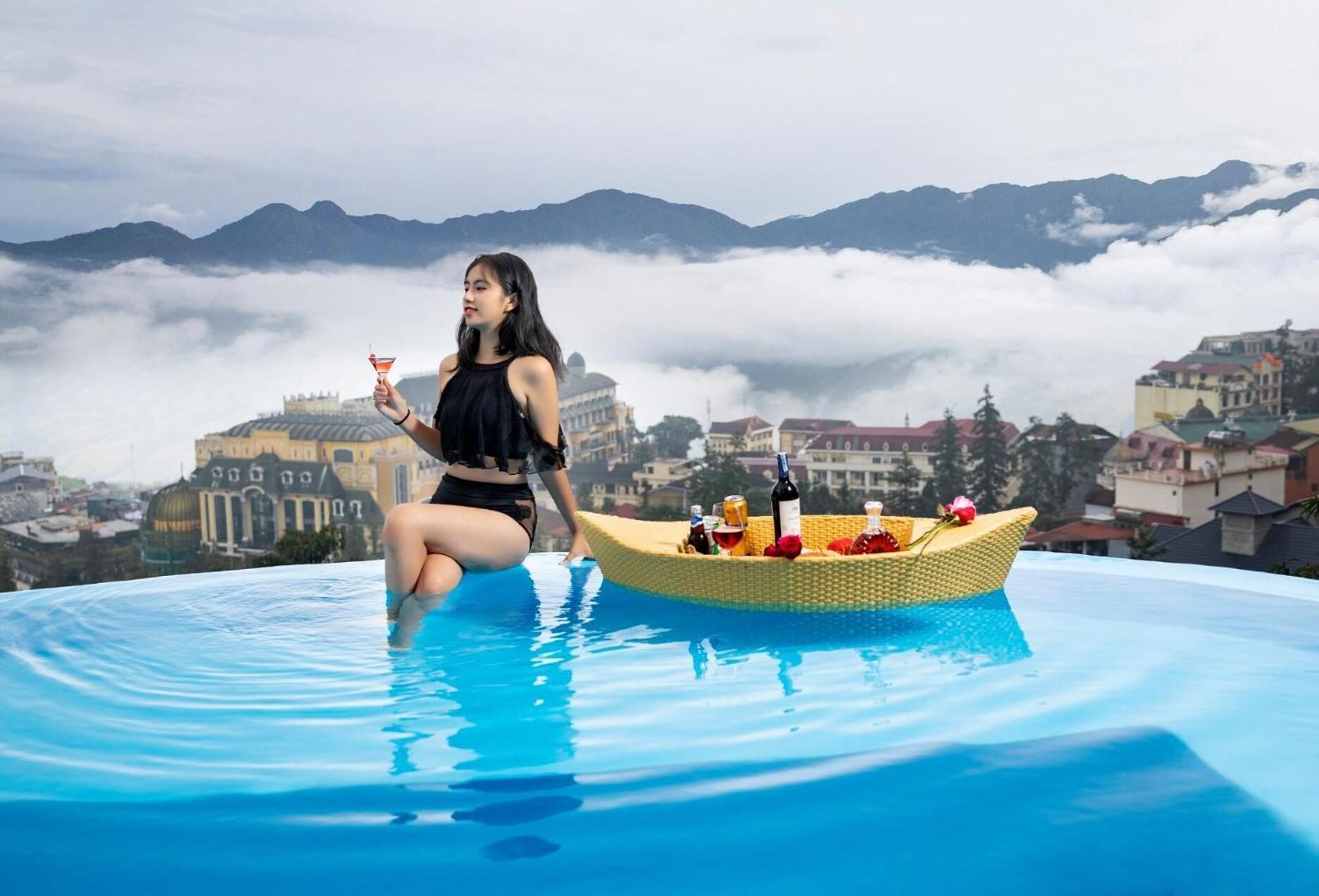 Una persona sentada al borde de una piscina, con un bote de mimbre que contiene botellas y una rosa.