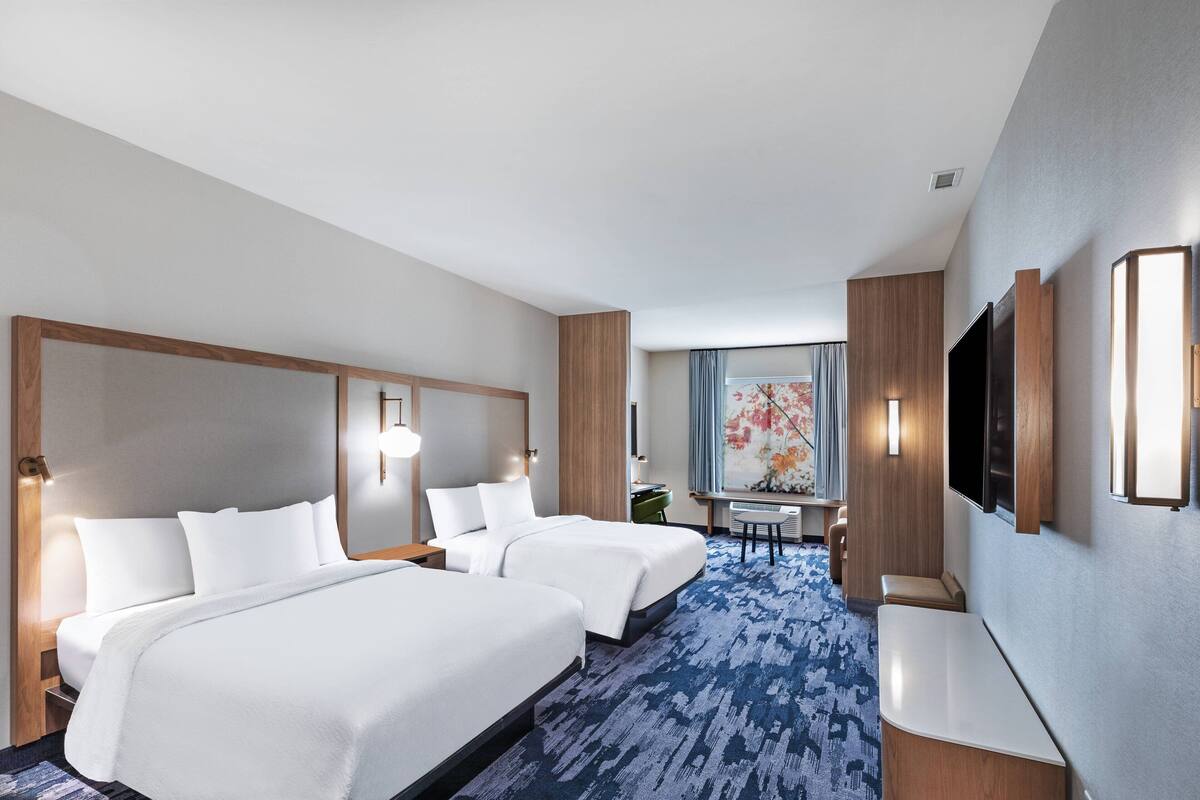A hotel room with two beds, a desk, a chair, a TV, and a large window with curtains.