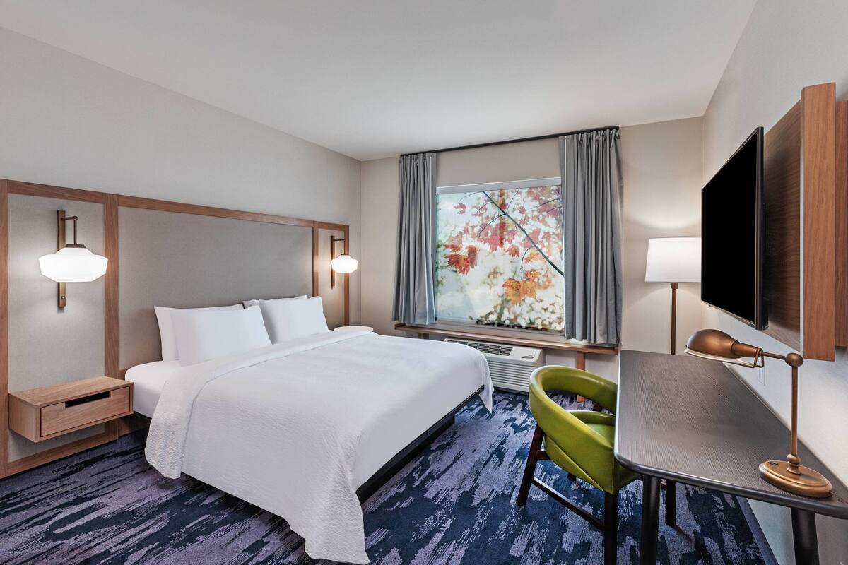A hotel room with a bed, a desk with a lamp, a chair, a large window with curtains, and a flat-screen TV.