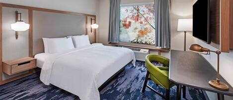 A hotel room with a bed, a desk with a lamp, a chair, a large window with curtains, and a flat-screen TV.