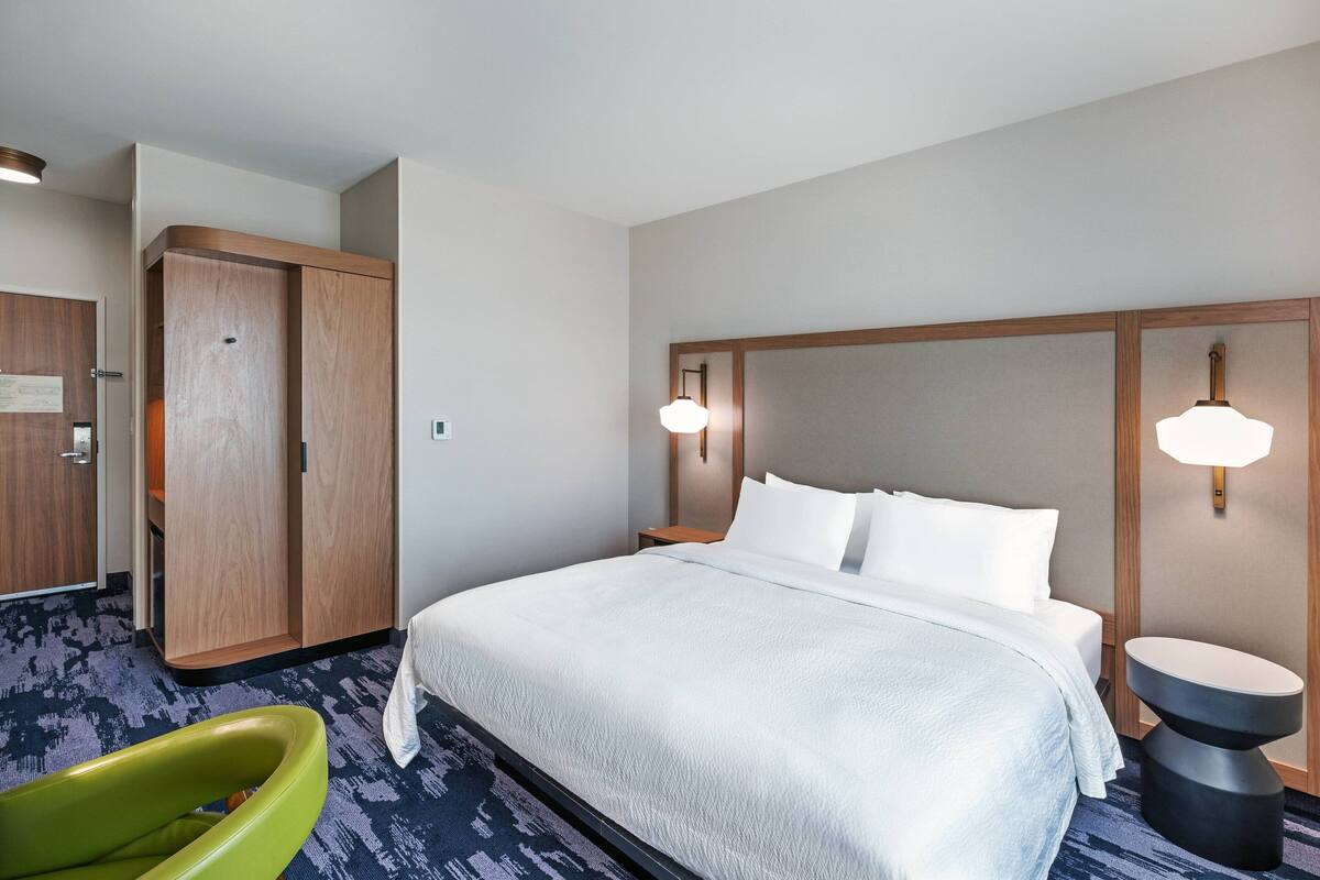 A hotel room with a large bed, a wooden wardrobe, a green chair, and a small table.